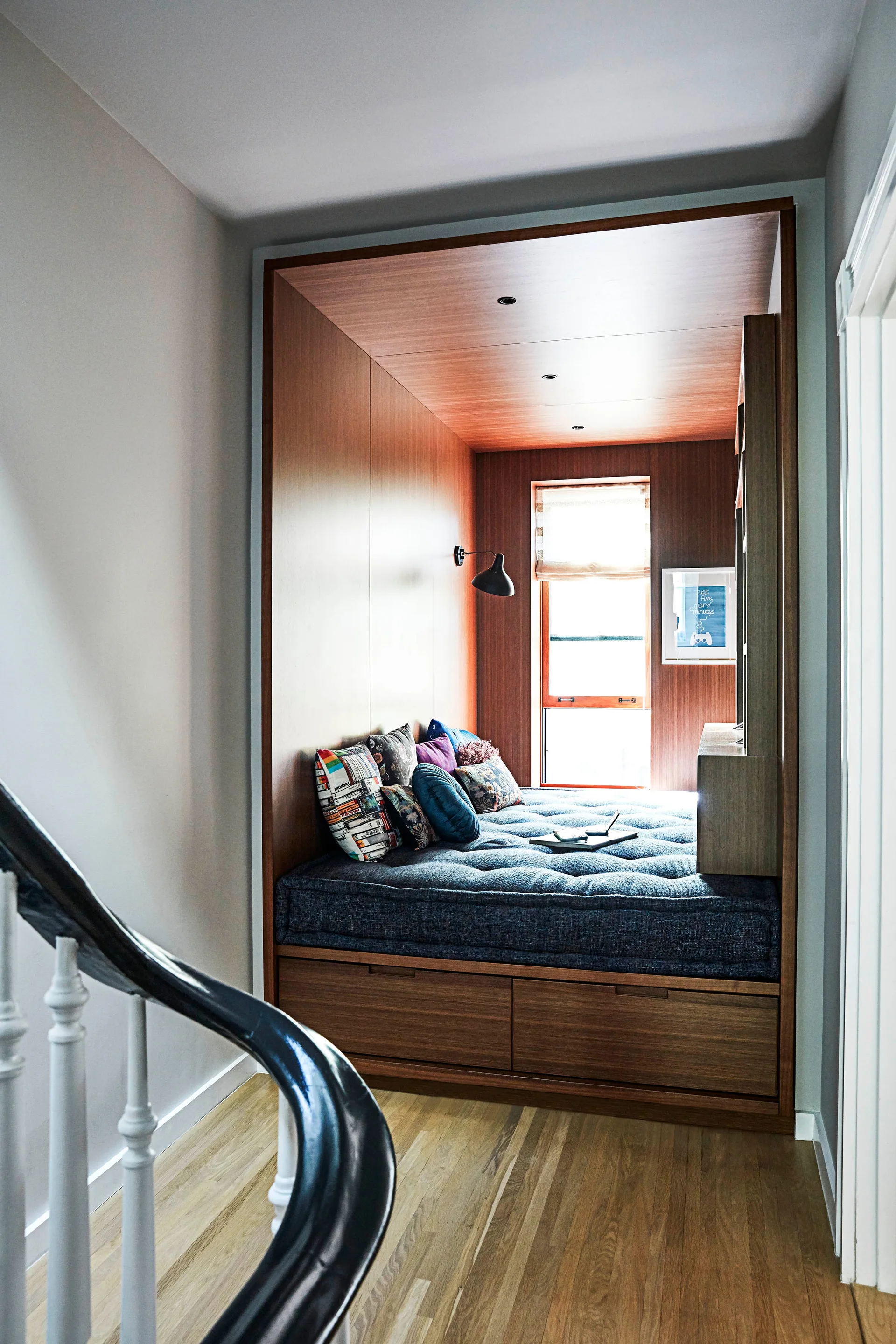 A built-in wooden sleeping alcove features a tufted blue daybed with decorative pillows, integrated under-bed drawers, and a black wall sconce.
