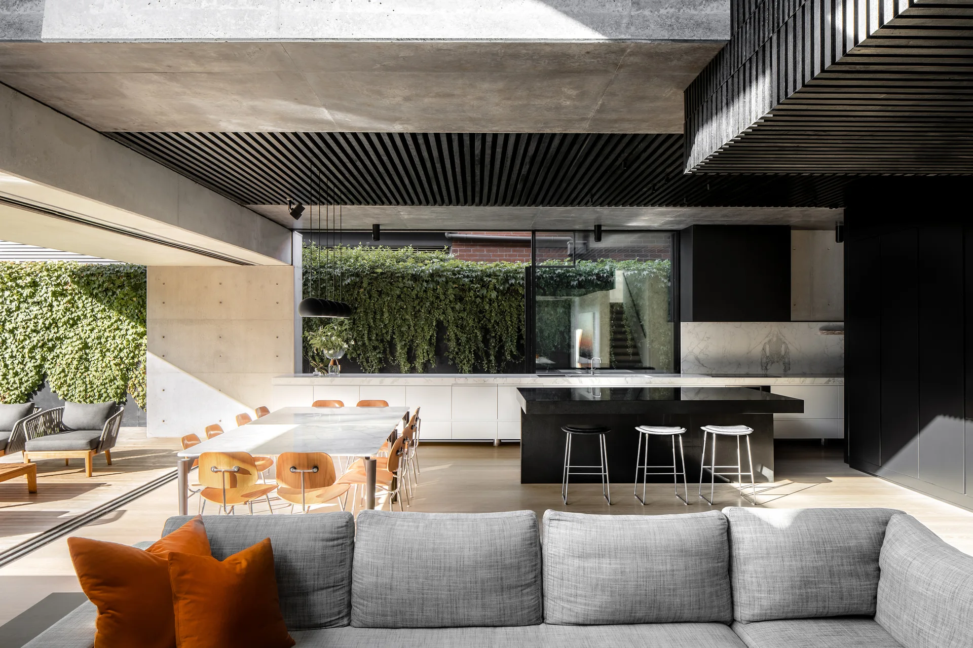 A modern open-concept kitchen and dining area features a black island, a marble dining table with wooden chairs, and a black slatted ceiling next to a terrace.