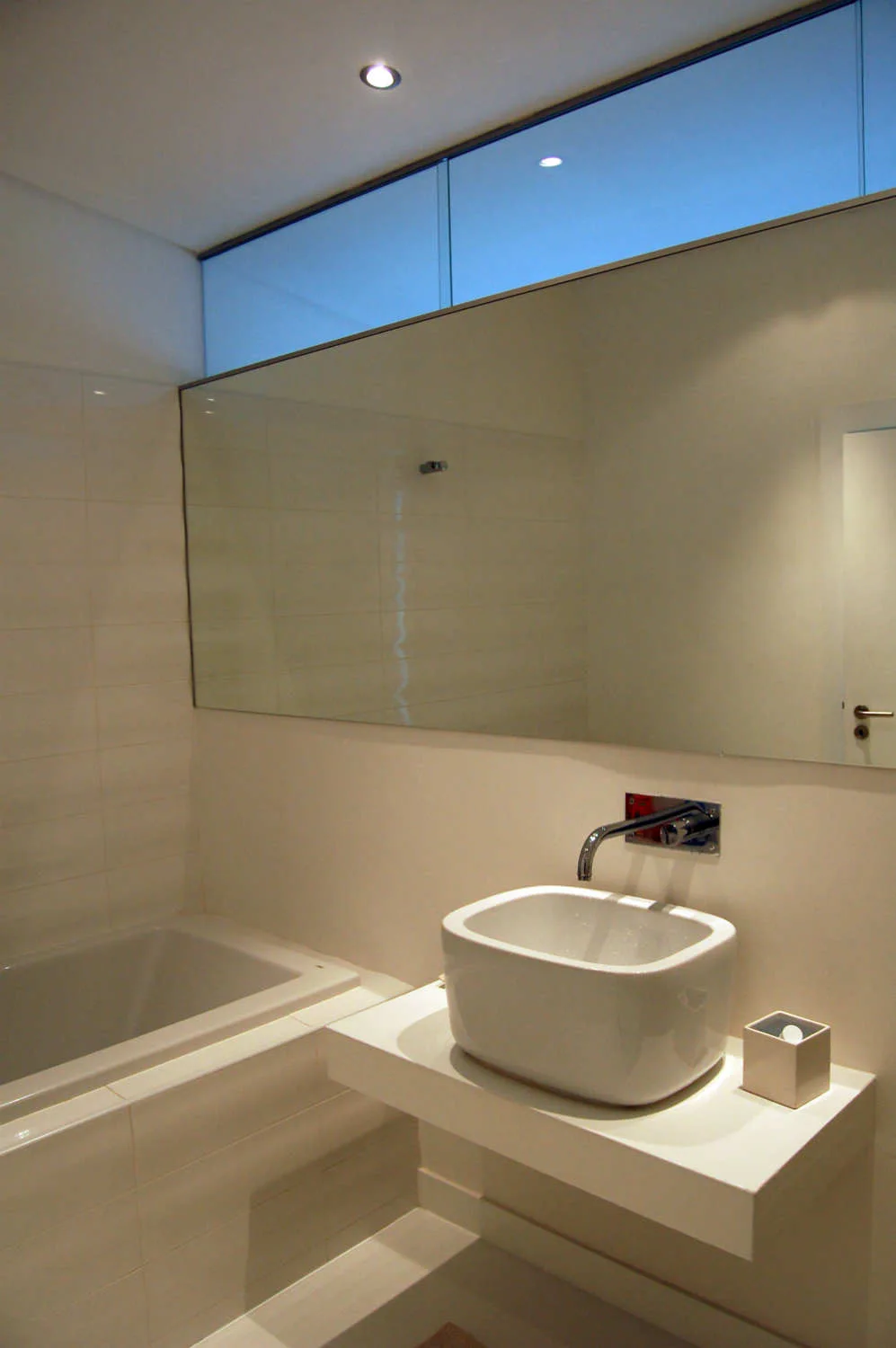 A minimalist white bathroom featuring a white vessel sink on a floating vanity console, a large wall mirror, and a high horizontal transom window.