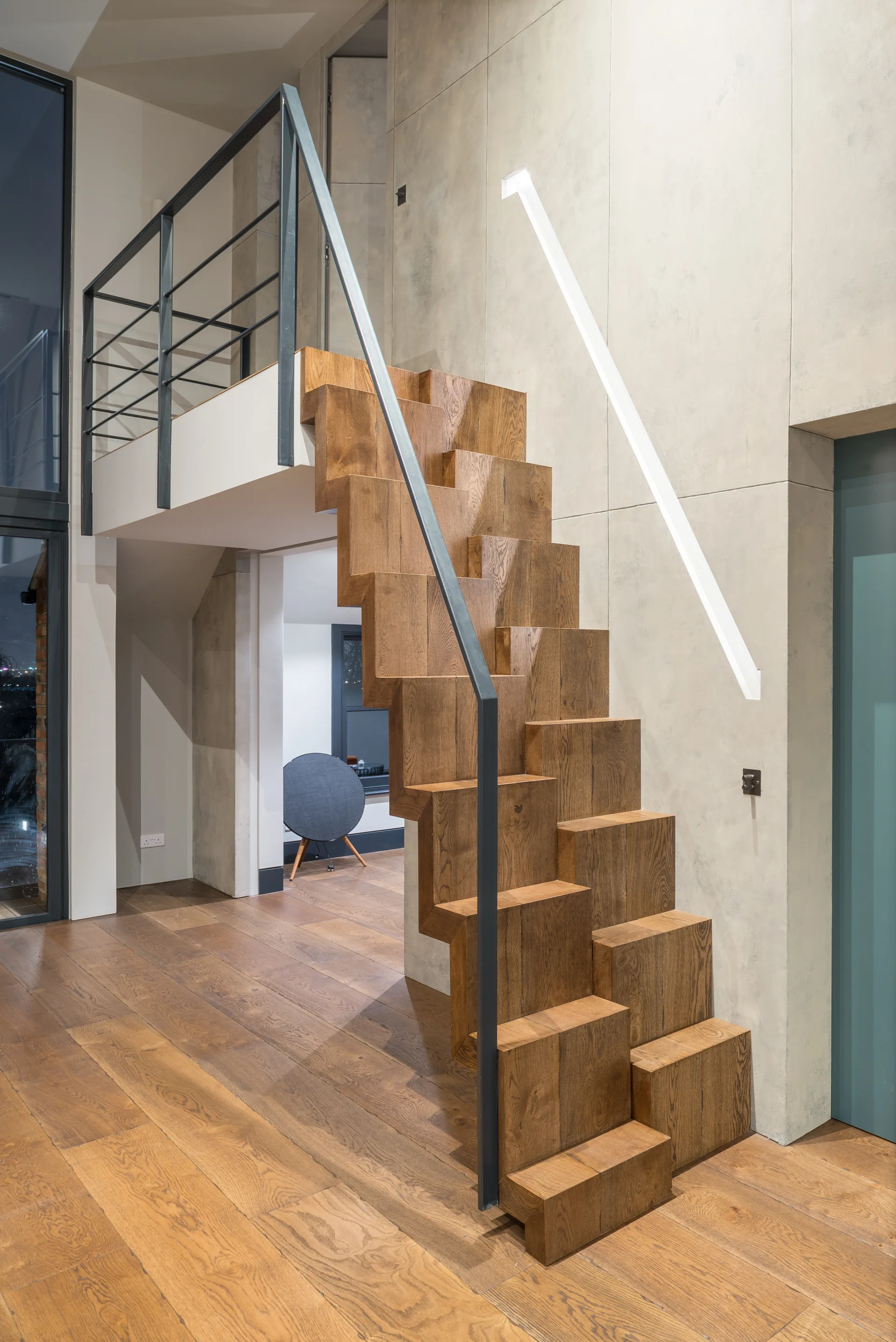 A modern wooden staircase with sculptural block steps features a simple black metal railing against a paneled greige wall.