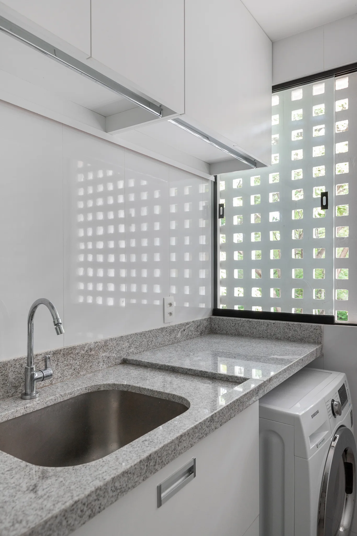 A compact white utility room features a speckled granite countertop with a sink, a washing machine, and a geometric patterned window screen.