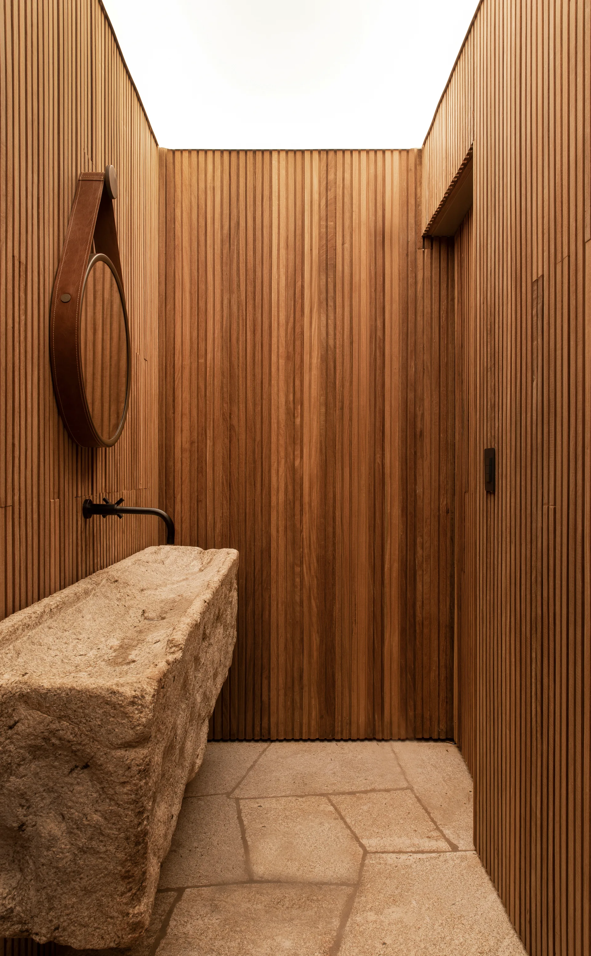 A minimalist powder room features vertical wood slat walls, a rough-hewn stone washbasin, a black faucet, and stone flooring.