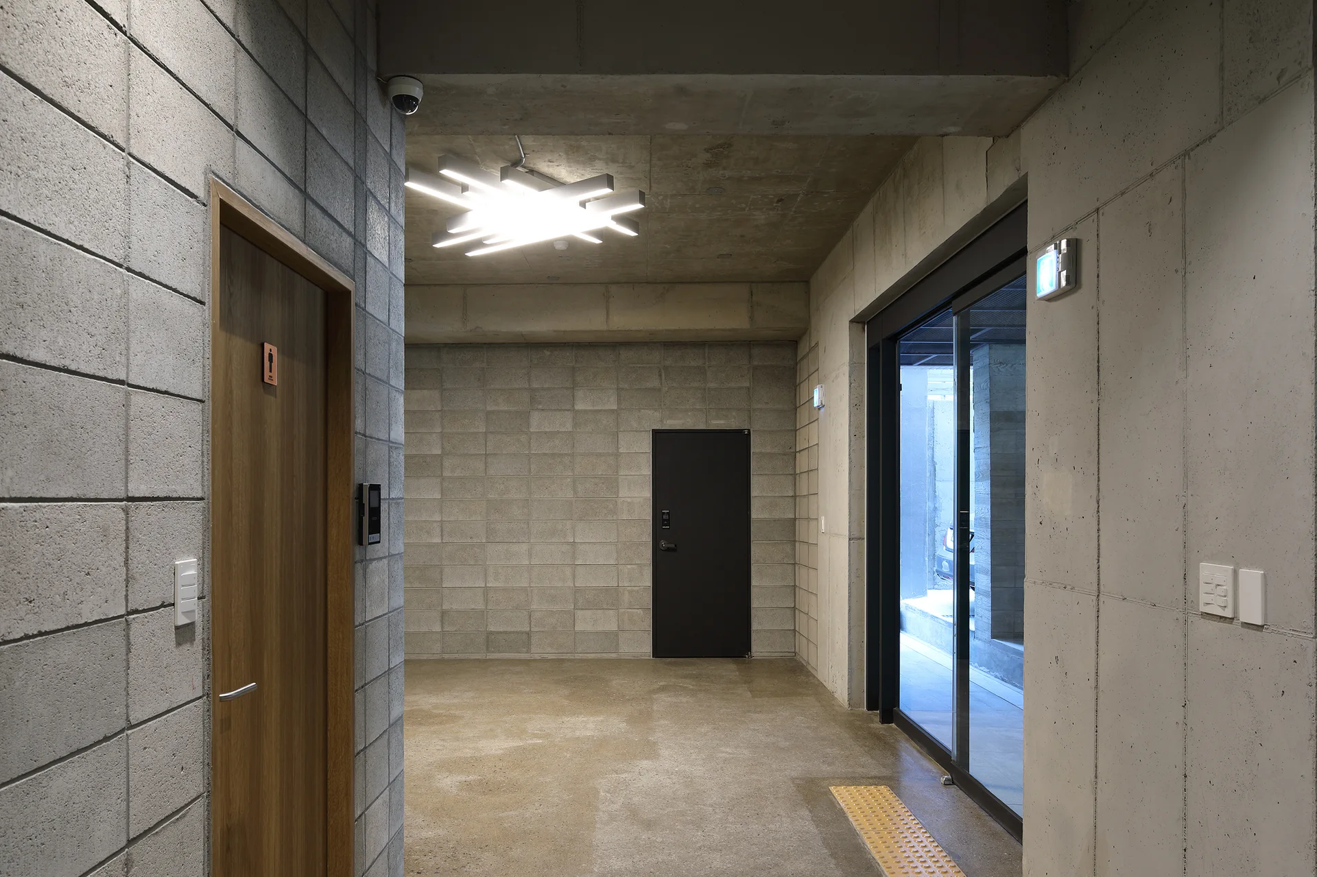 A minimalist hallway features exposed concrete block walls, a polished concrete floor, a geometric ceiling light, and a warm wooden door.