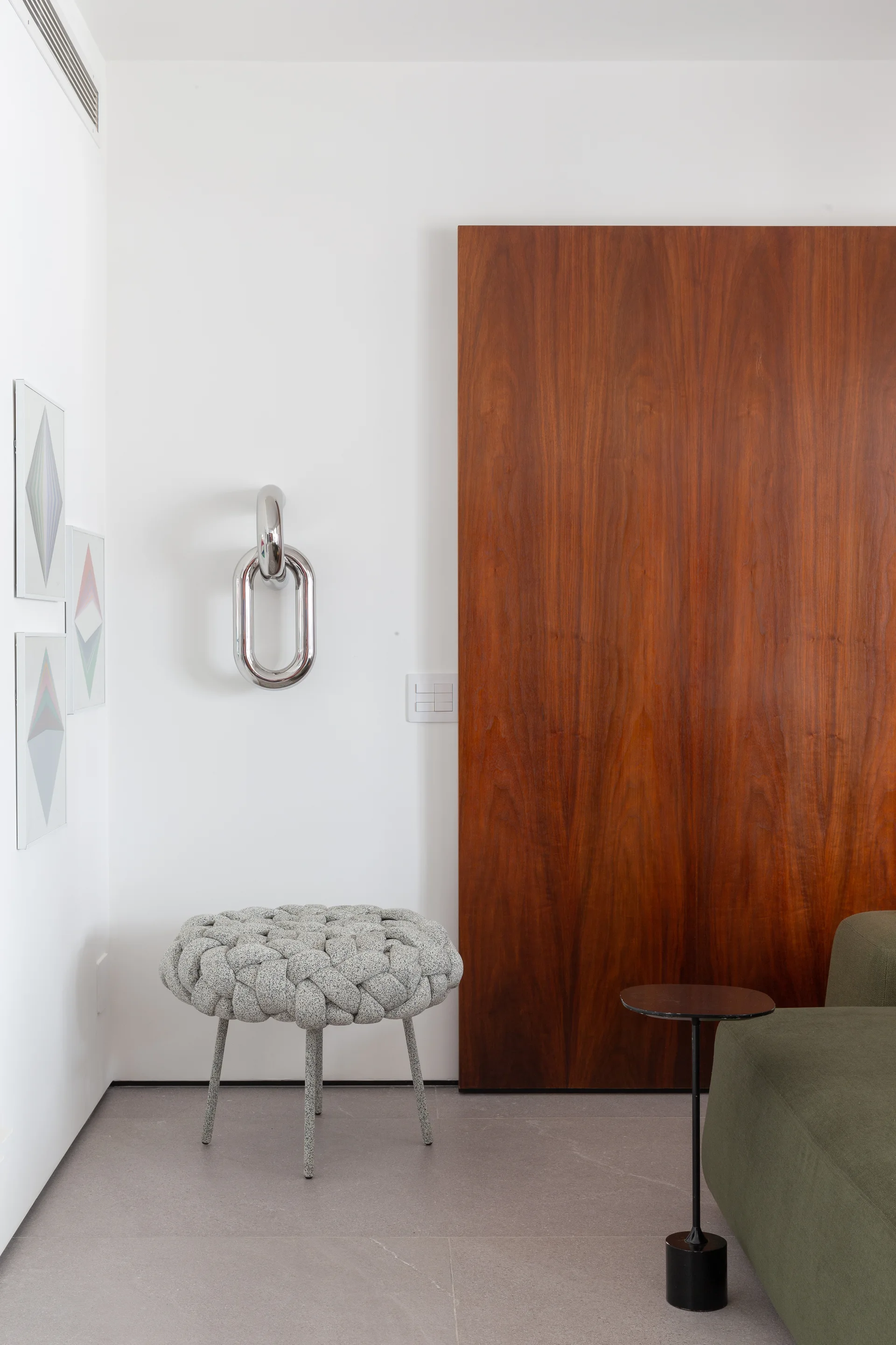 A minimalist room features a grey woven stool, a polished chrome chain sculpture on a white wall, and a large dark wood sliding panel.