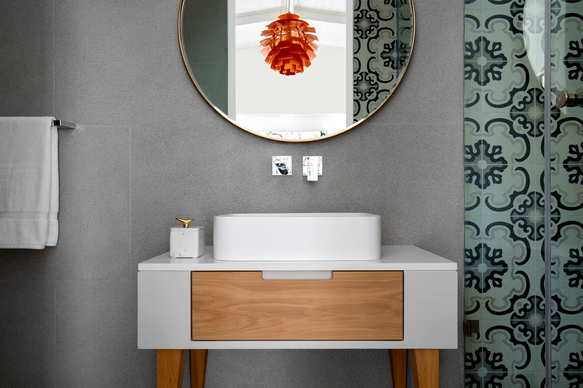 A modern bathroom features a white and wood vanity with a vessel sink, a round brass mirror, and grey tiled walls.