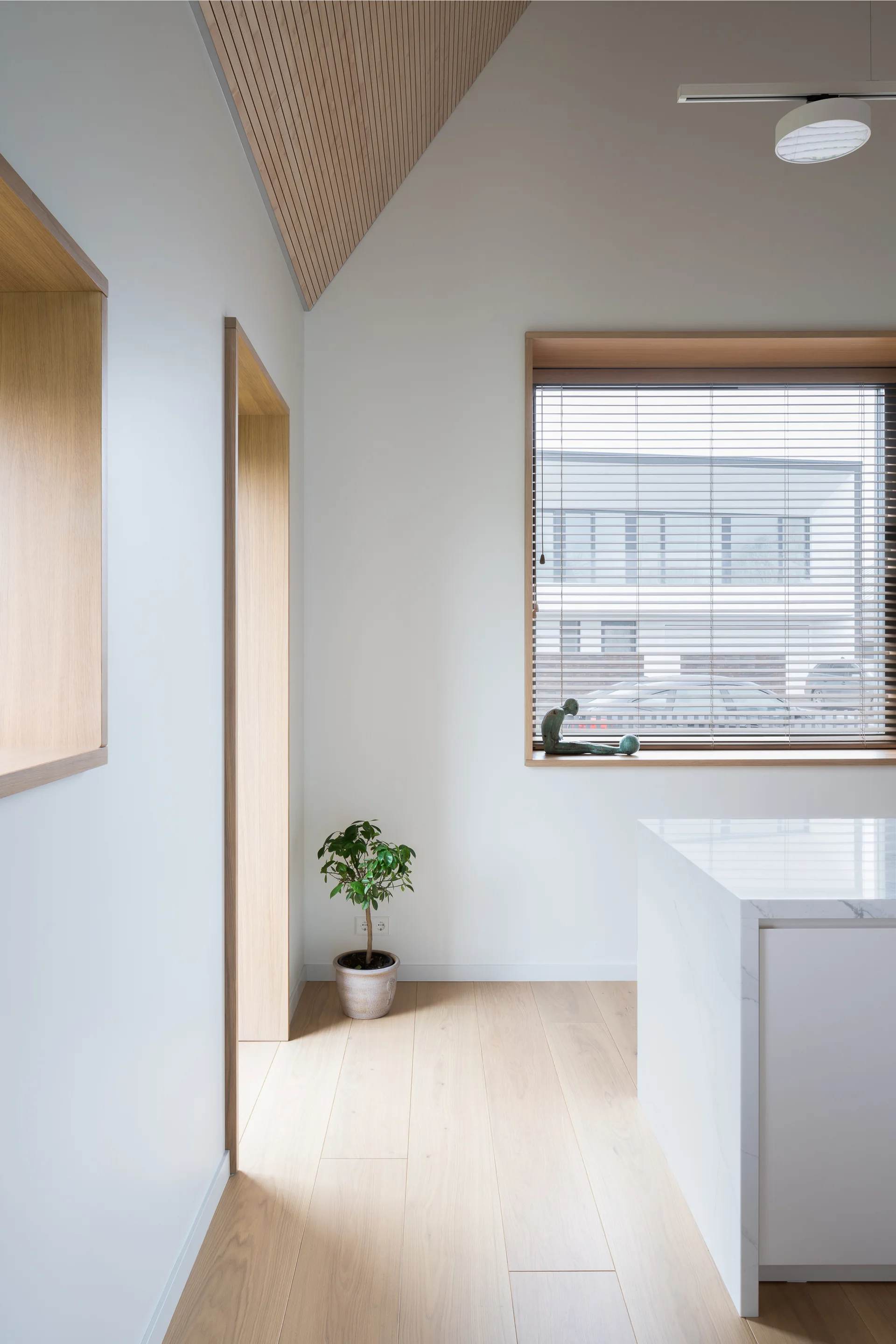 A minimalist kitchen space featuring a white marble island, a sloped wood-slat ceiling, and light oak floorboards alongside a large window.