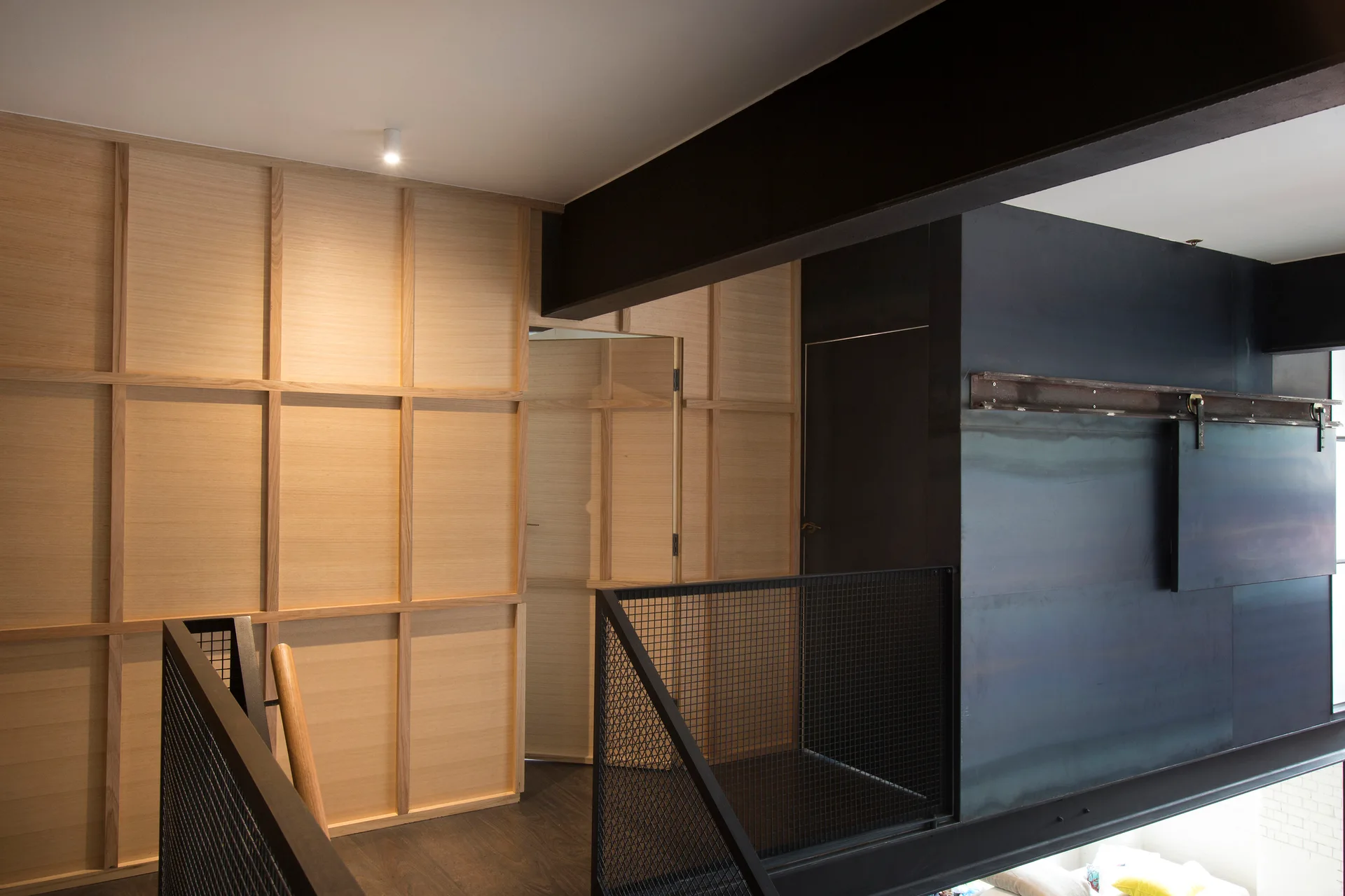 An upper-level loft landing featuring a light wood grid wall panel, black metal mesh railings, and a heavy black steel structural beam.