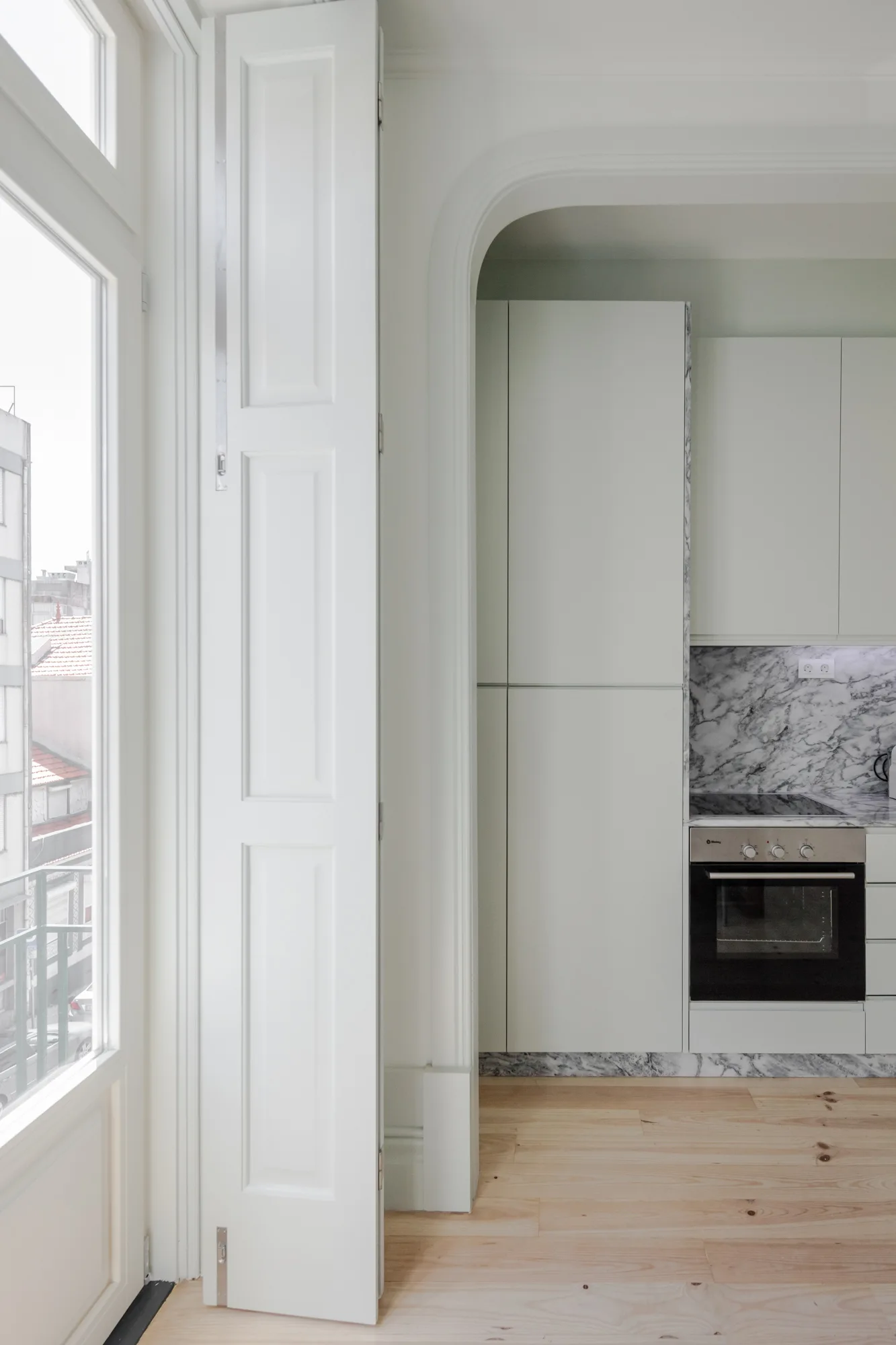 A view through a white curved archway into a minimalist kitchen with light gray flat-panel cabinets, a veined marble backsplash, and light wood floors.