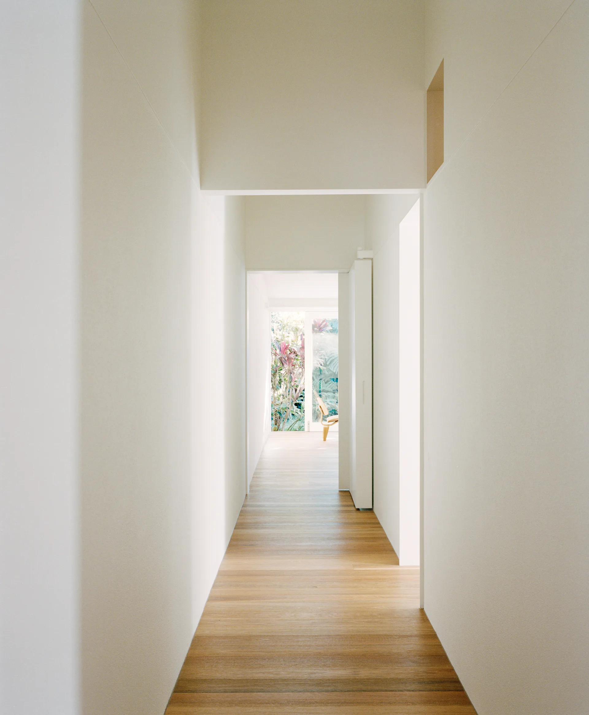 A long minimalist hallway with white walls and wood plank floors leading to a sunlit room with green foliage.