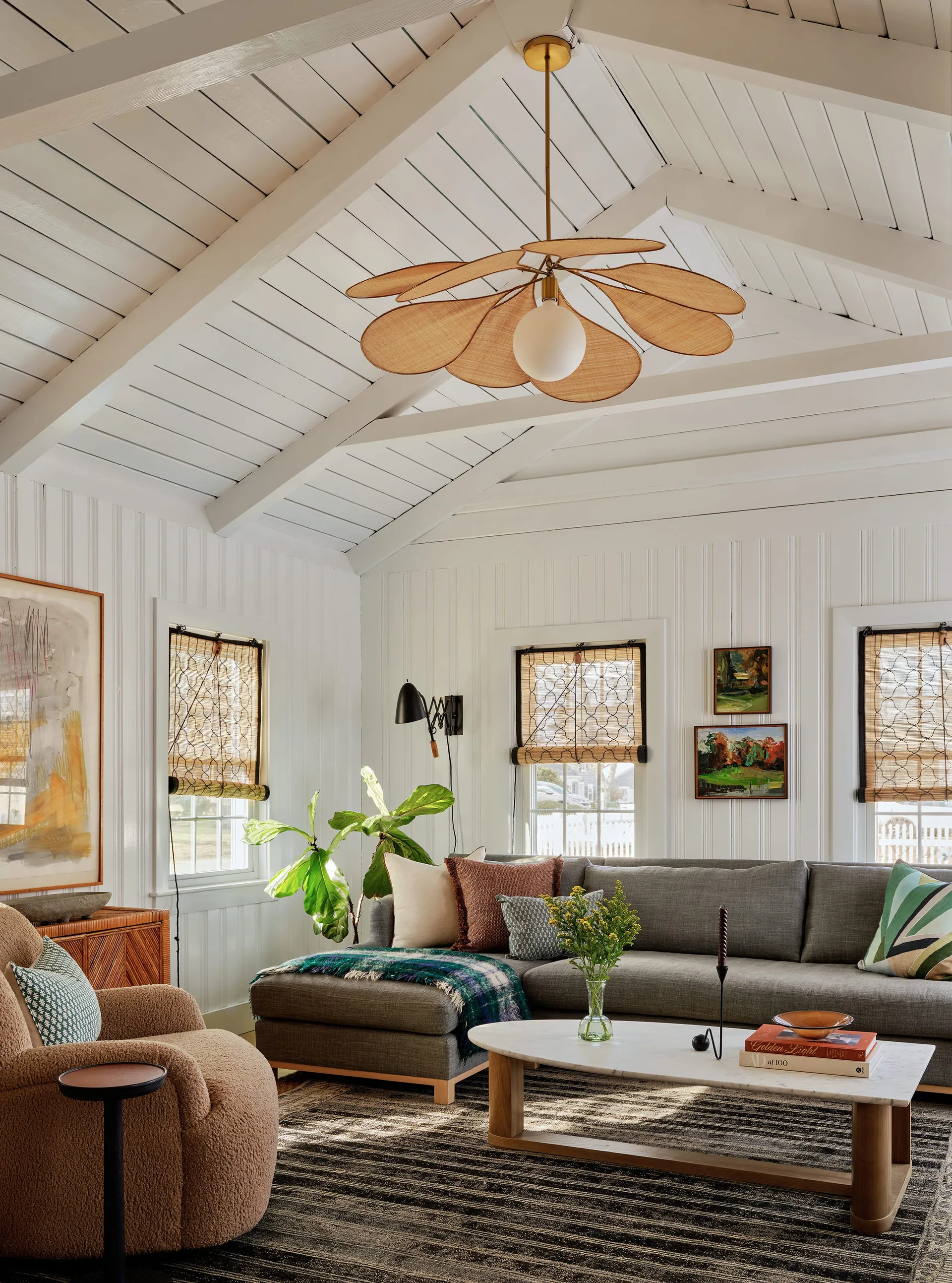 A light-filled living room featuring a grey sectional, a textured tan armchair, and a unique petal-shaped rattan chandelier under a vaulted ceiling.