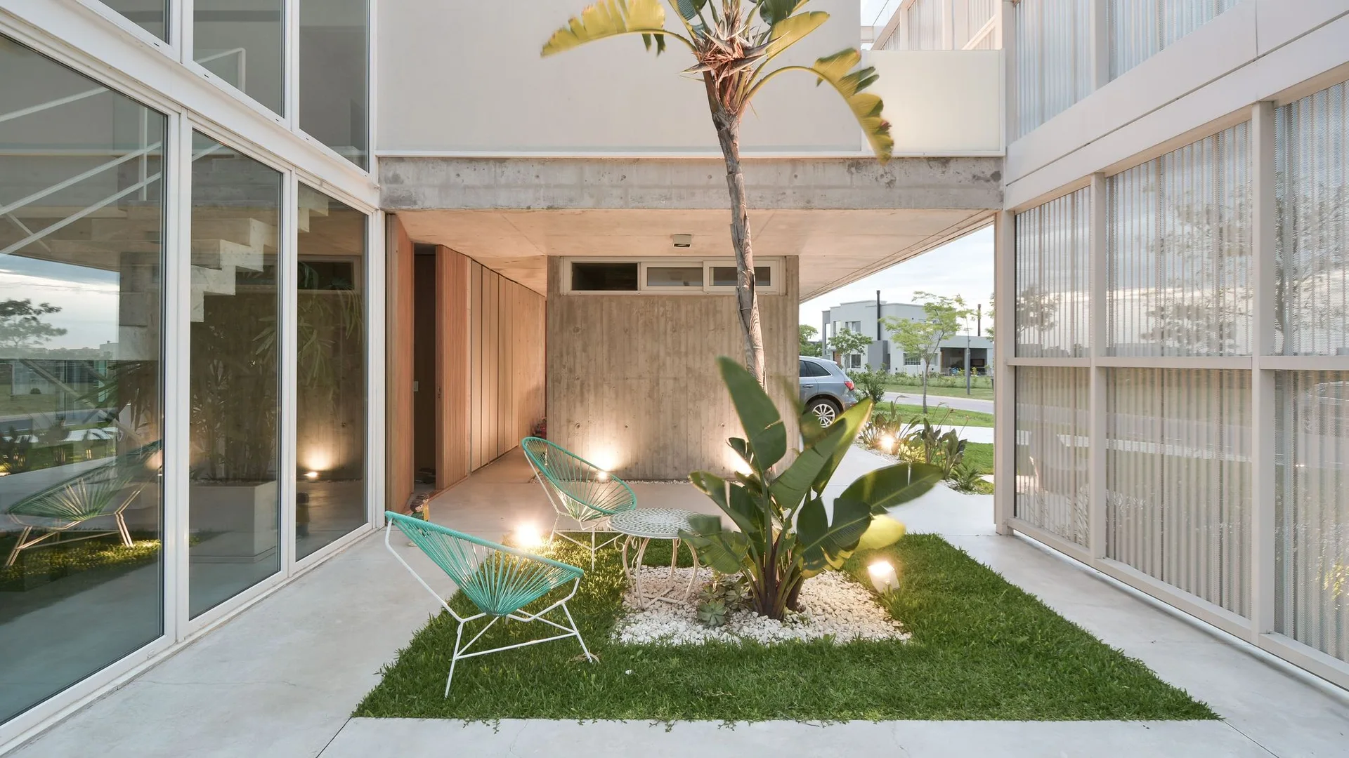 Two teal wire chairs sit on a grass patch within a concrete courtyard next to a palm tree and glass-walled modern house.