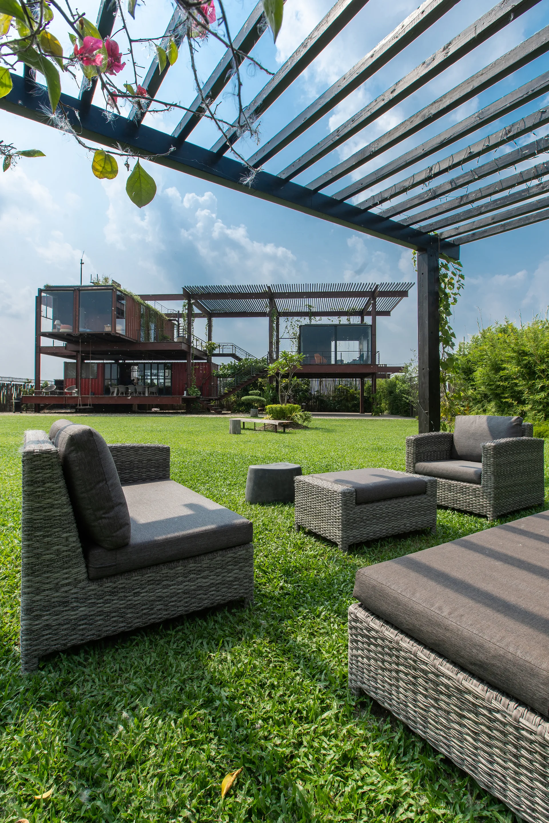 A gray wicker lounge set with plush cushions is arranged on a green lawn beneath a black slatted pergola next to a modern metal structure.