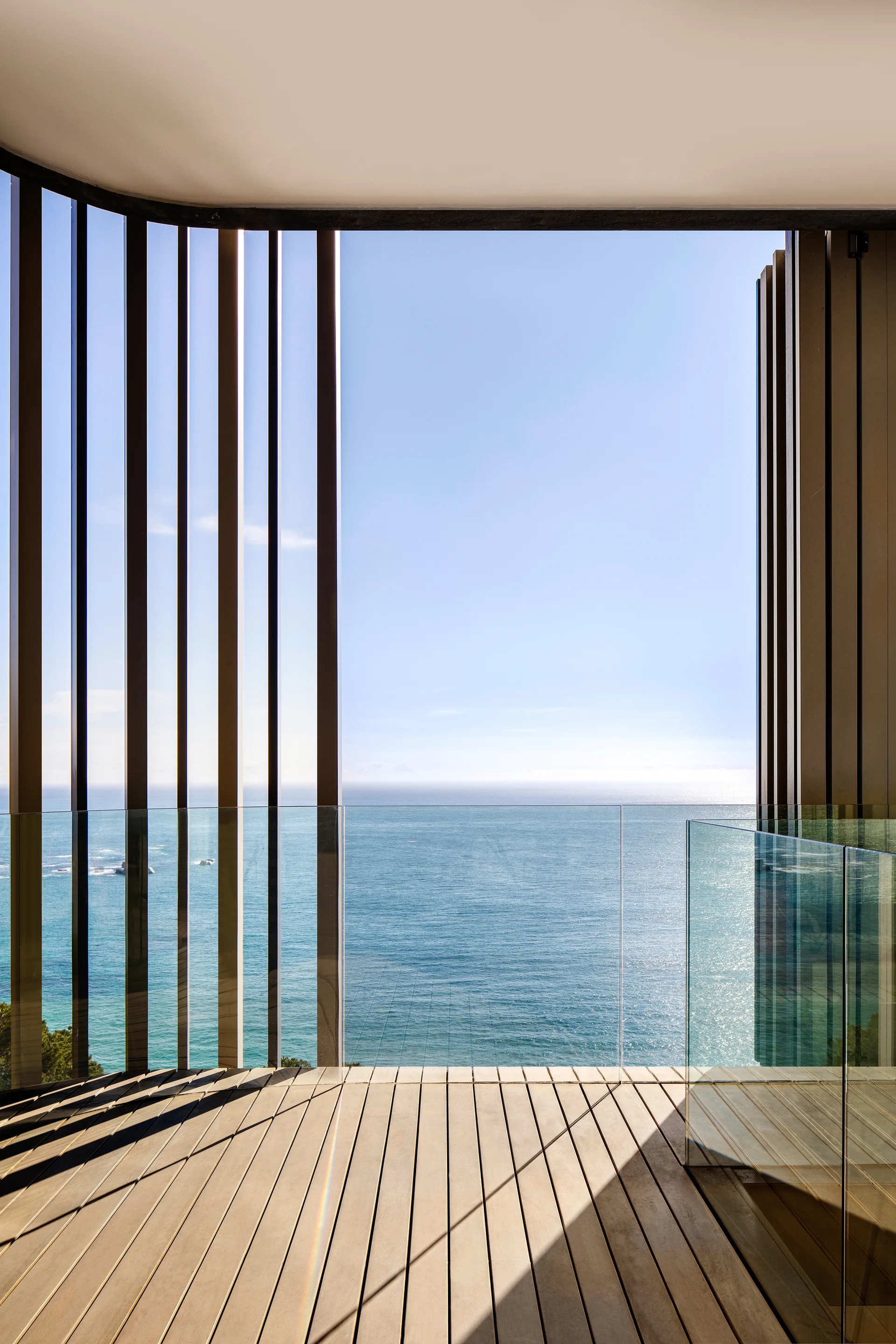 A modern outdoor balcony with light wood decking, a clear glass railing, and tall vertical architectural slats overlooking the ocean.