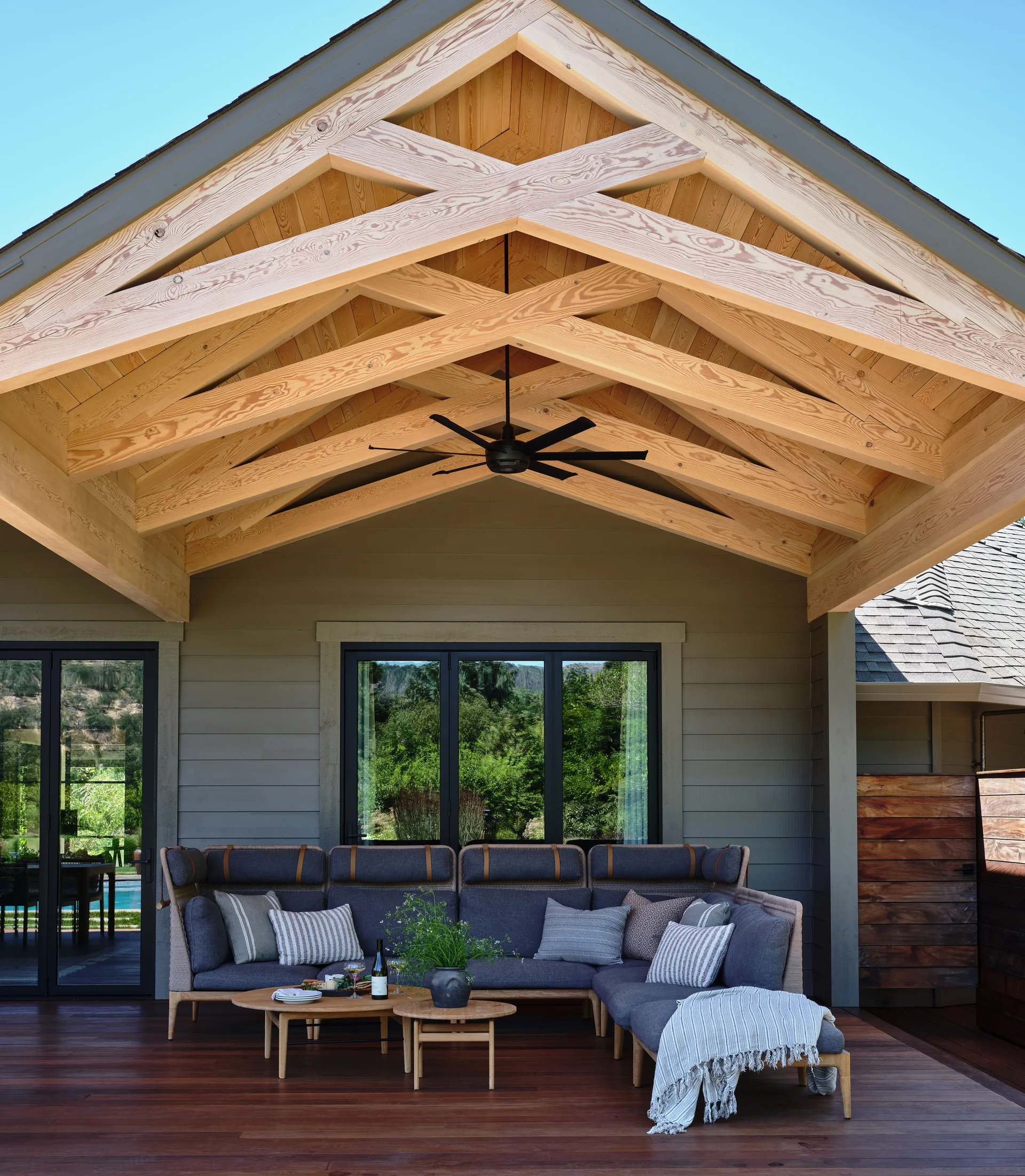 A grey outdoor sectional sofa and wooden nesting tables sit on a dark wood deck beneath a vaulted timber truss ceiling.