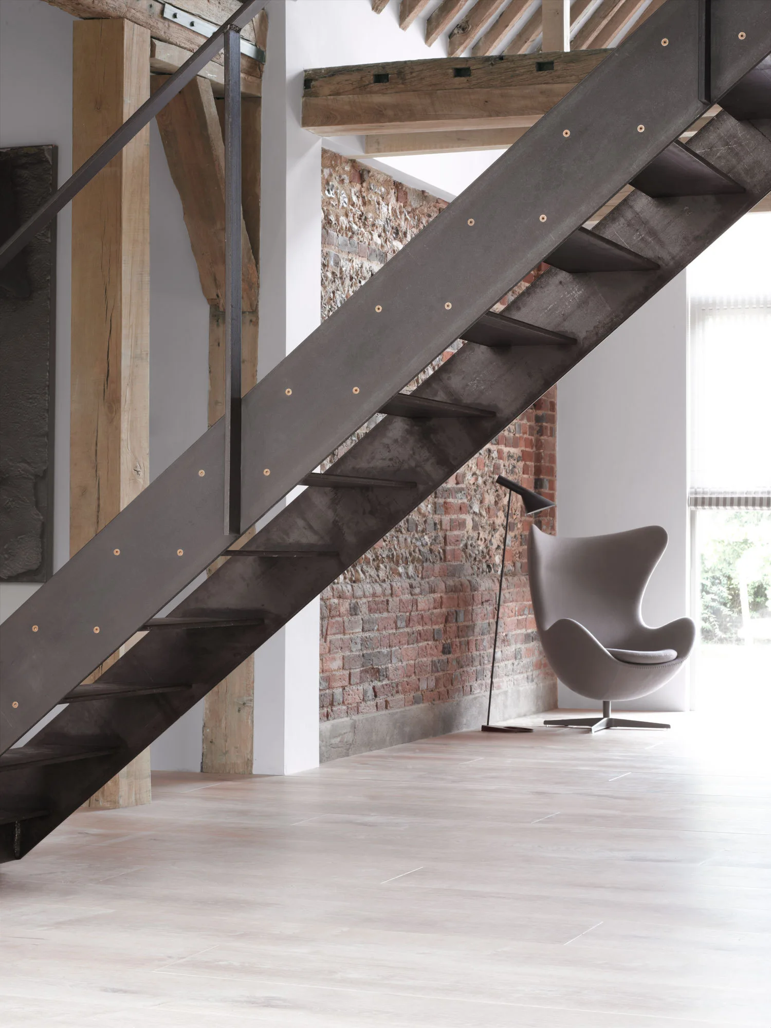 An industrial dark metal staircase stands next to an exposed brick wall, featuring an accent chair and timber ceiling beams.