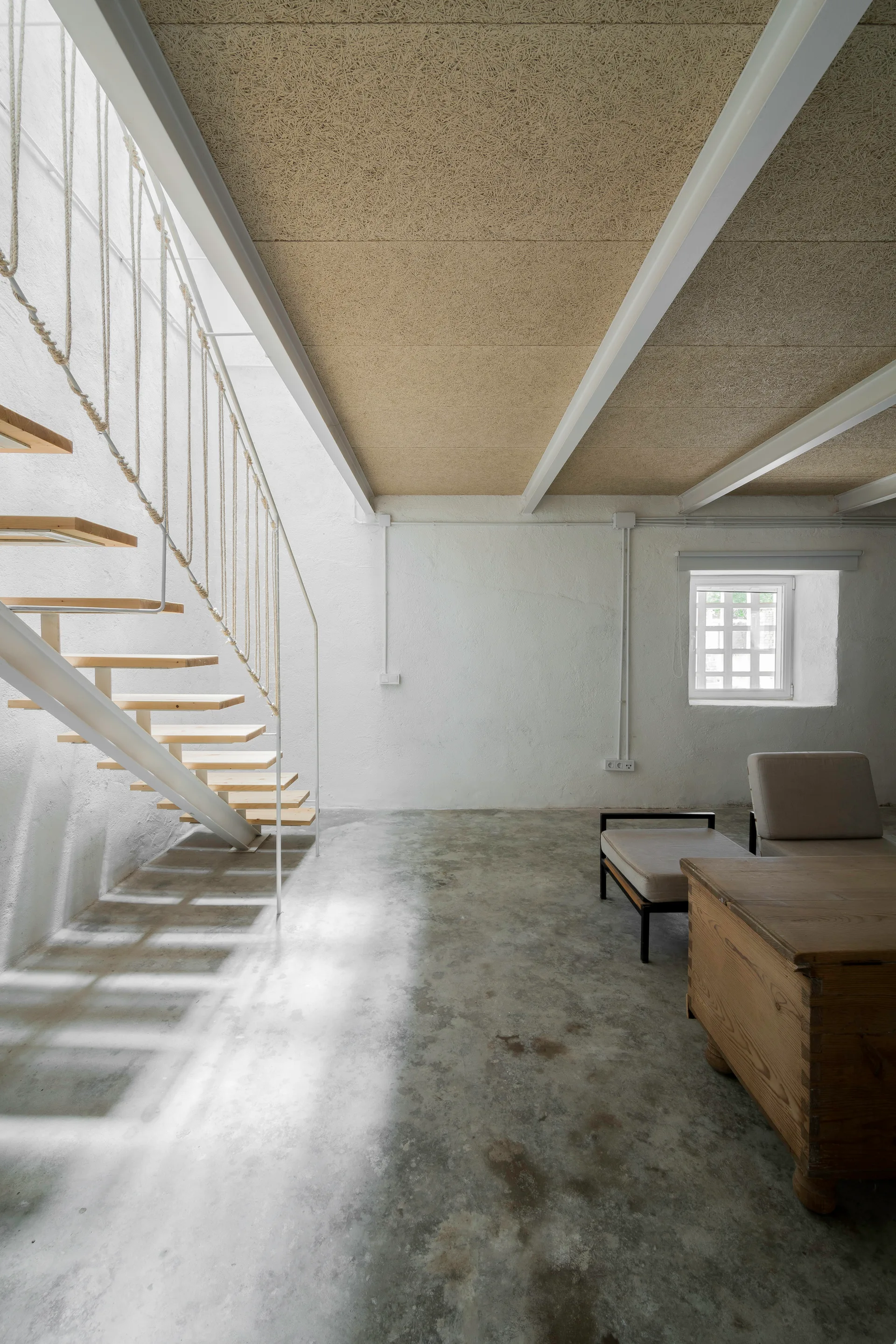 An industrial living room features an open-riser staircase with a white rope railing next to concrete flooring and a textured ceiling.