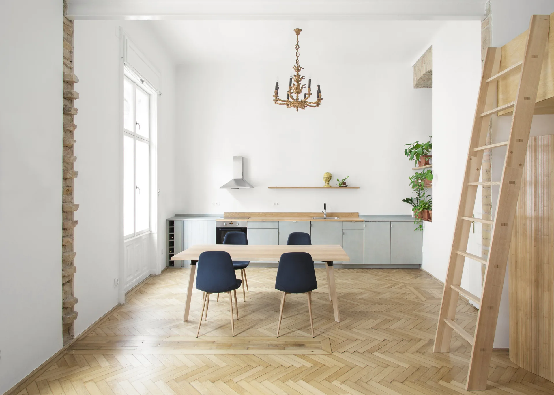An open kitchen and dining area features a light wood table with navy chairs, grey cabinetry, a tall wooden ladder, and herringbone parquet floors.
