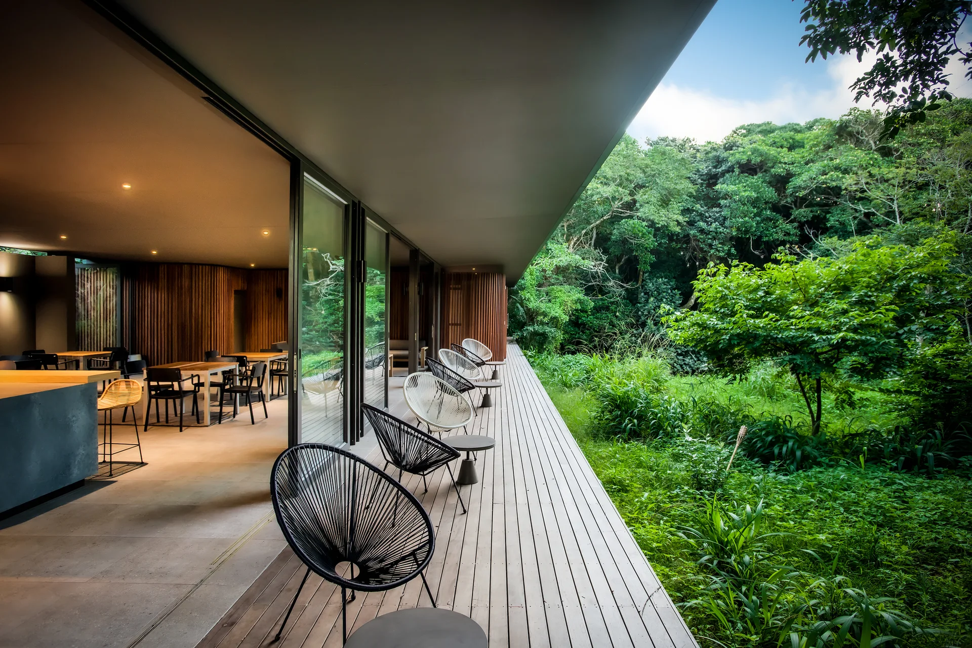 A long wooden terrace deck with black and white woven lounge chairs next to glass walls overlooking a lush green forest.