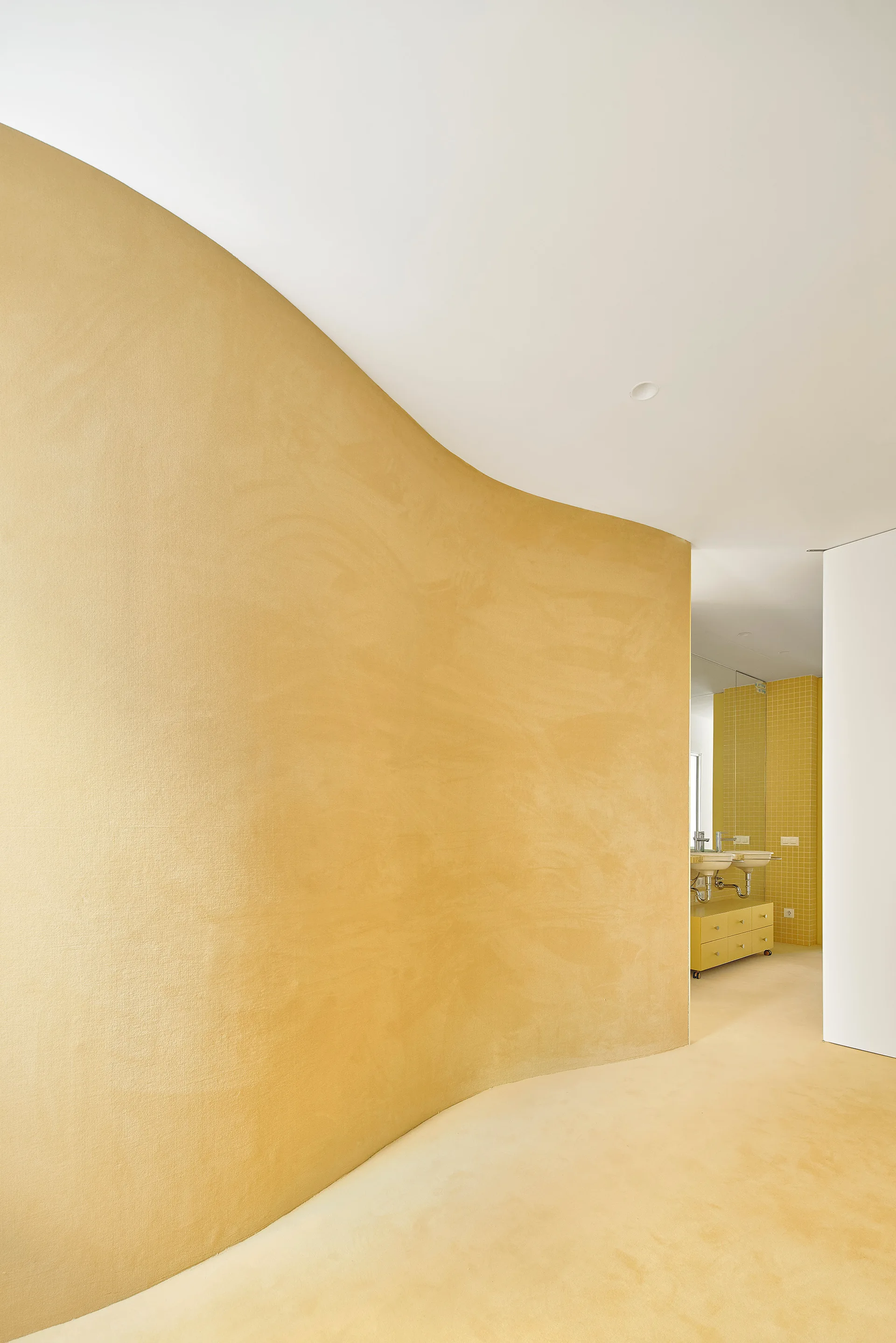 A minimalist bathroom interior features a sweeping curved yellow plaster wall that transitions into a tiled vanity area with double sinks.