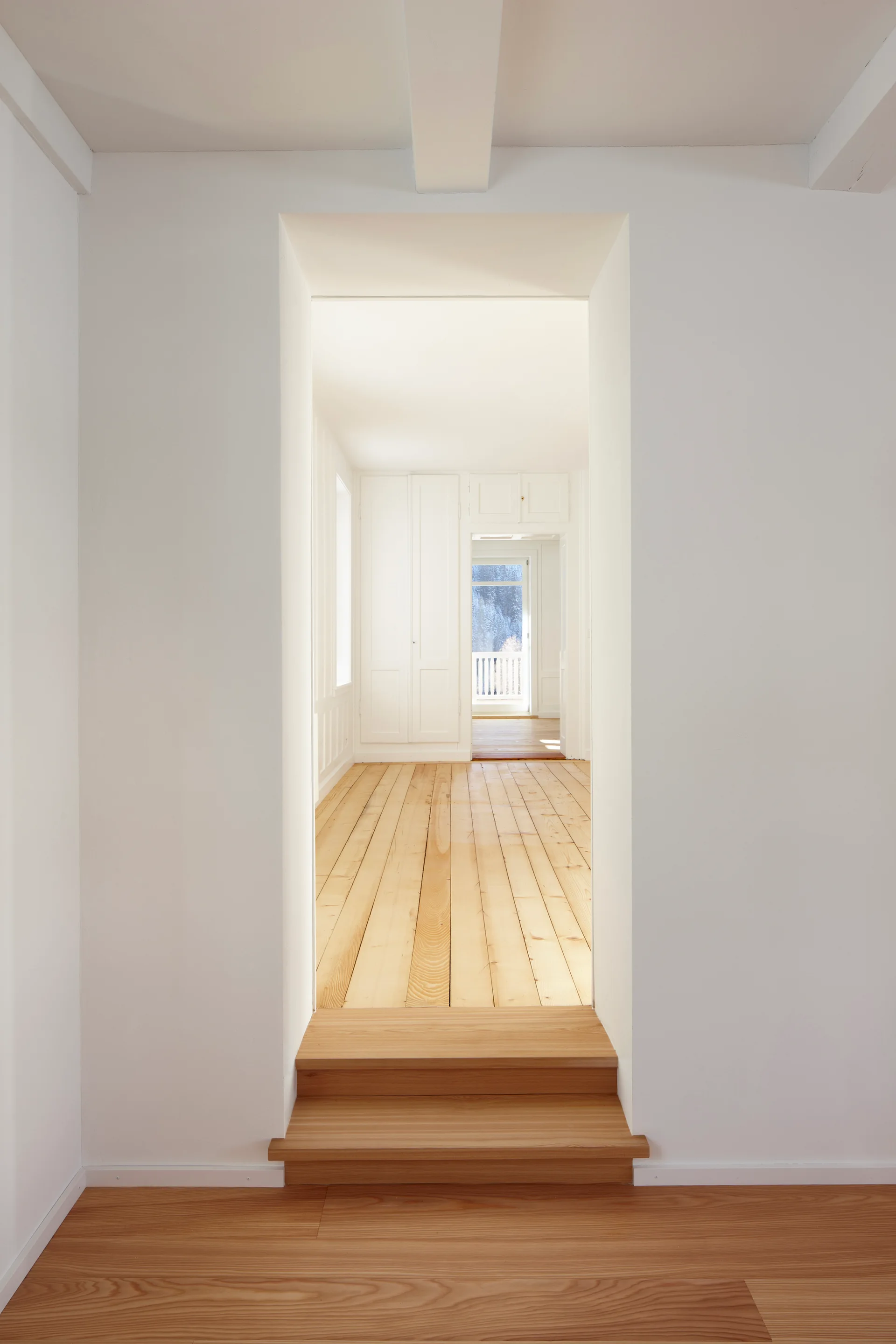 A deep rectangular doorway with two light wood steps leads into a minimalist hallway featuring matching wood plank flooring and white walls.