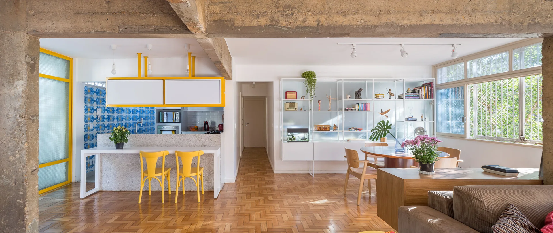 An open-plan living and kitchen area featuring parquet flooring, an exposed concrete beam, and a kitchen counter with yellow chairs.