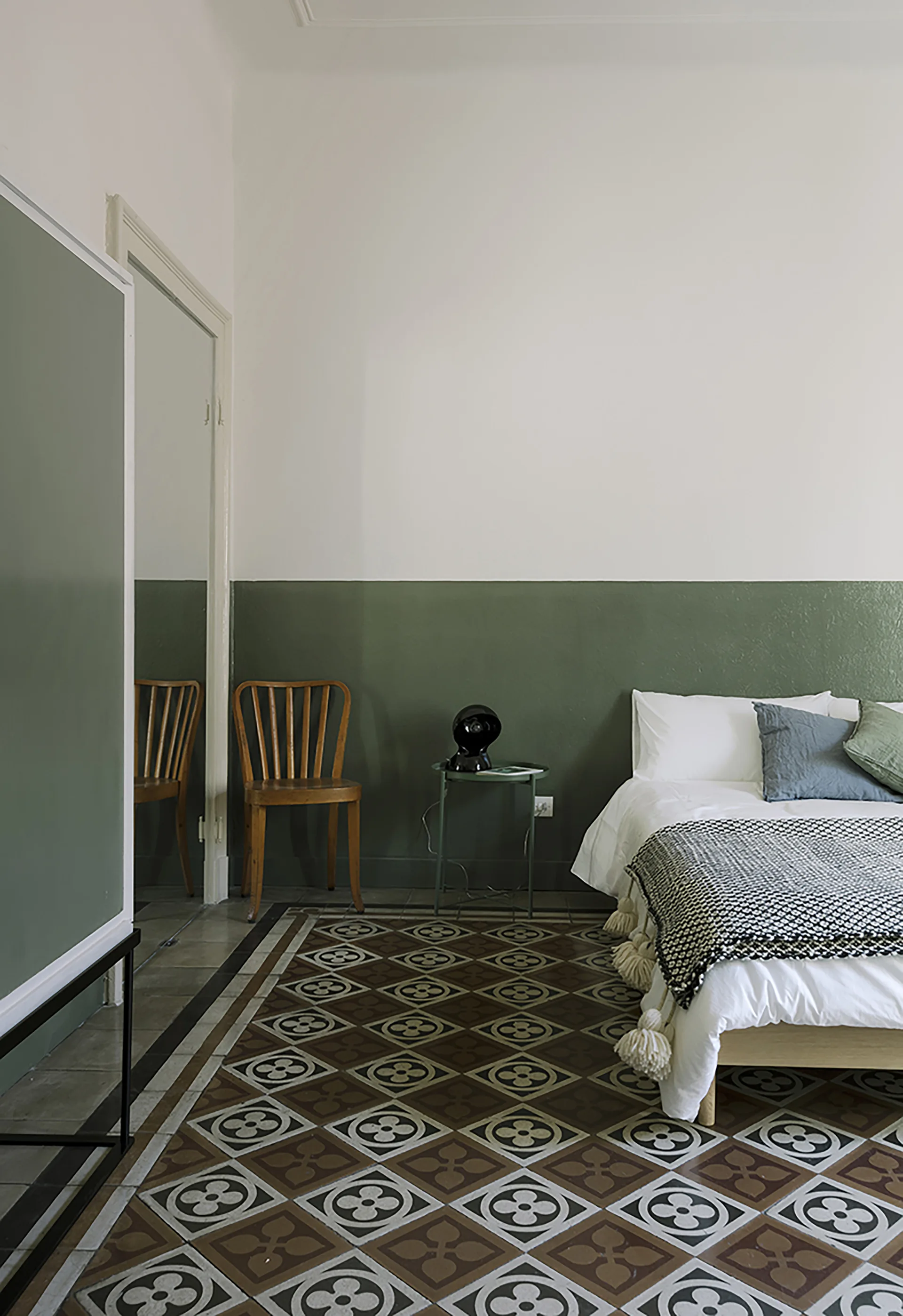A minimalist bedroom featuring a sage green painted half-wall, a platform bed with white bedding, and geometric patterned floor tiles.