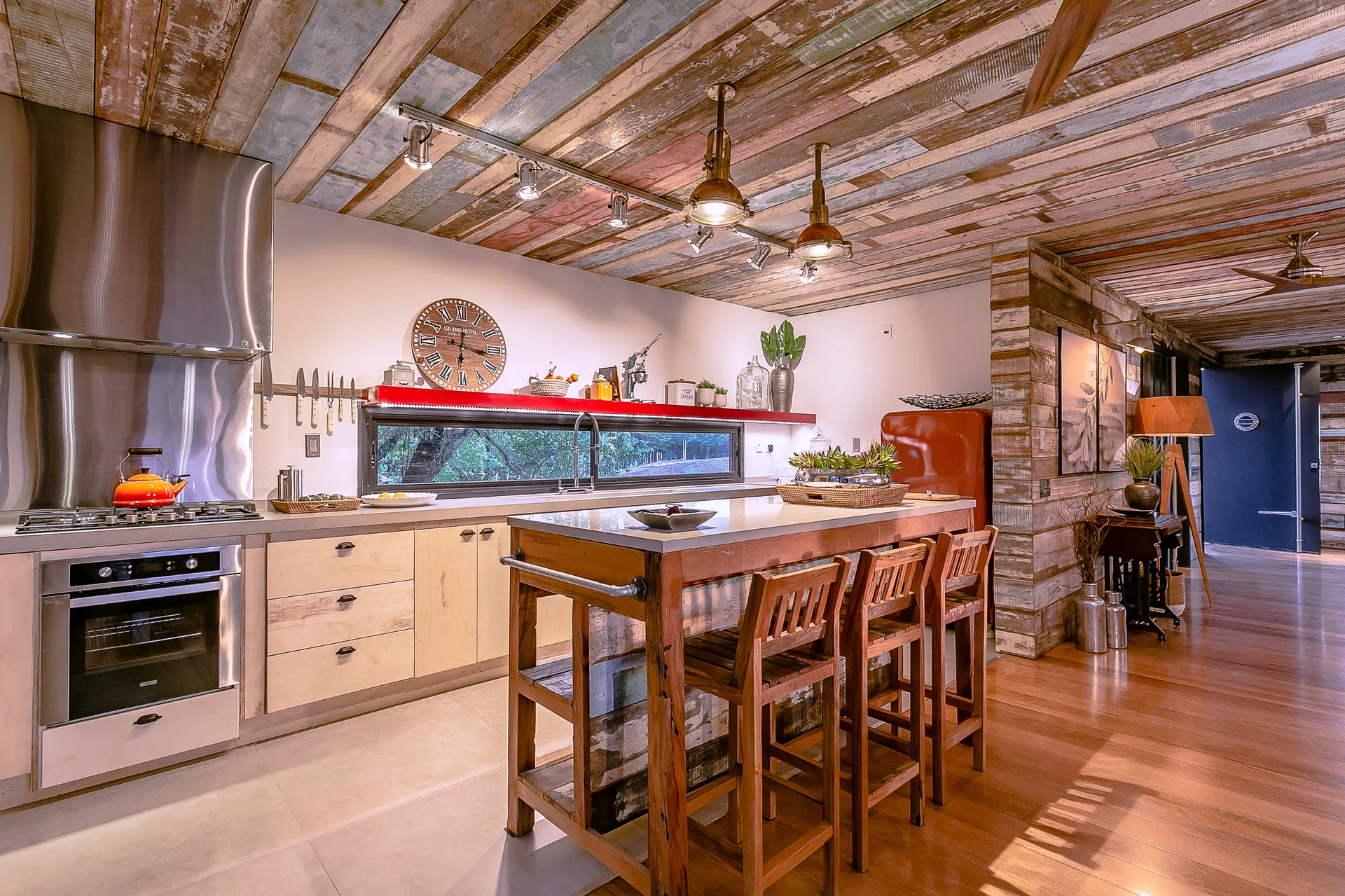 A rustic kitchen features a multi-toned reclaimed wood ceiling, a central wooden island with bar stools, and a long horizontal window.