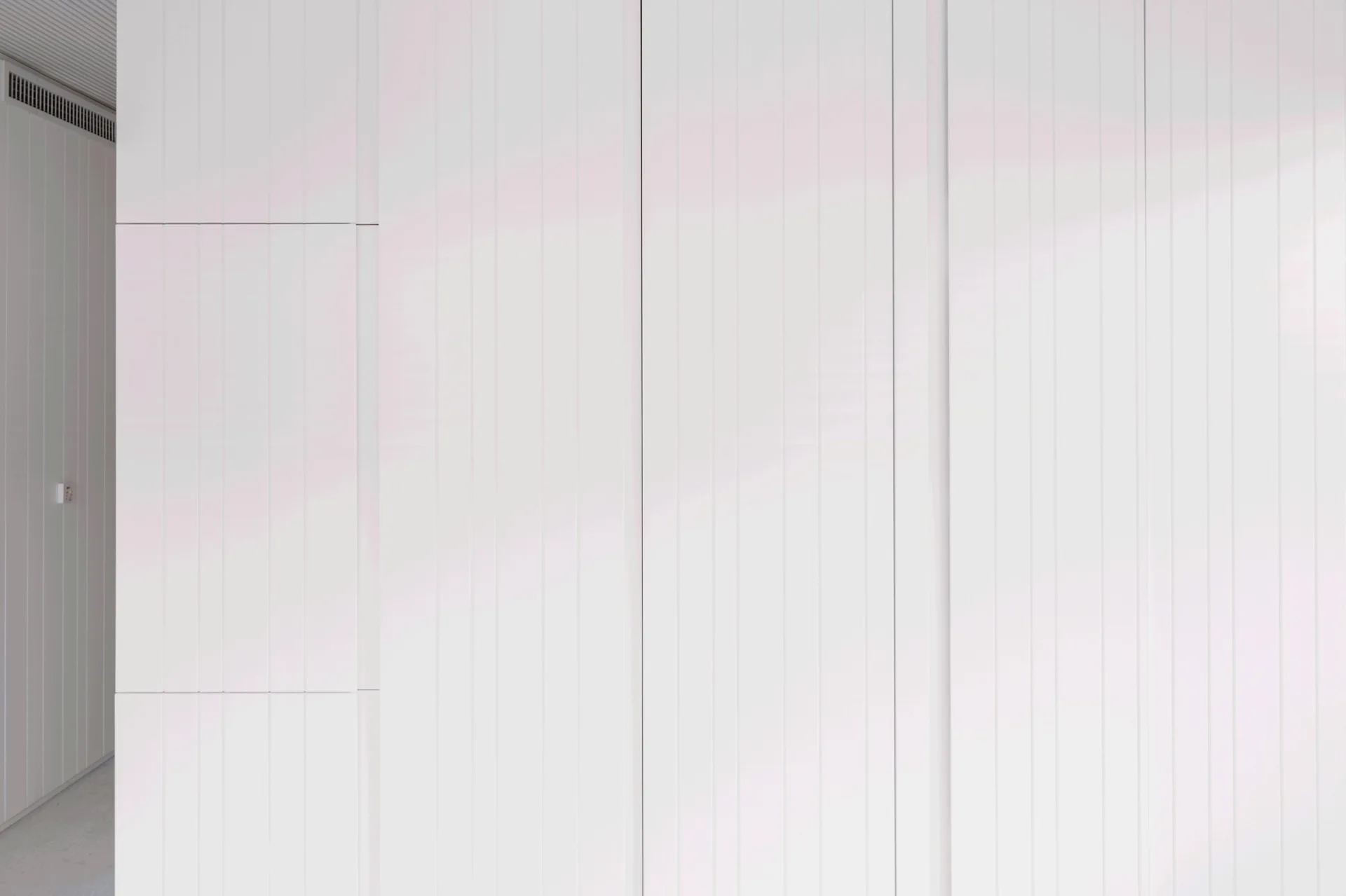 A minimalist white wall features floor-to-ceiling vertical paneling with thin groove lines, a hidden door seam, and an integrated vent at the top.