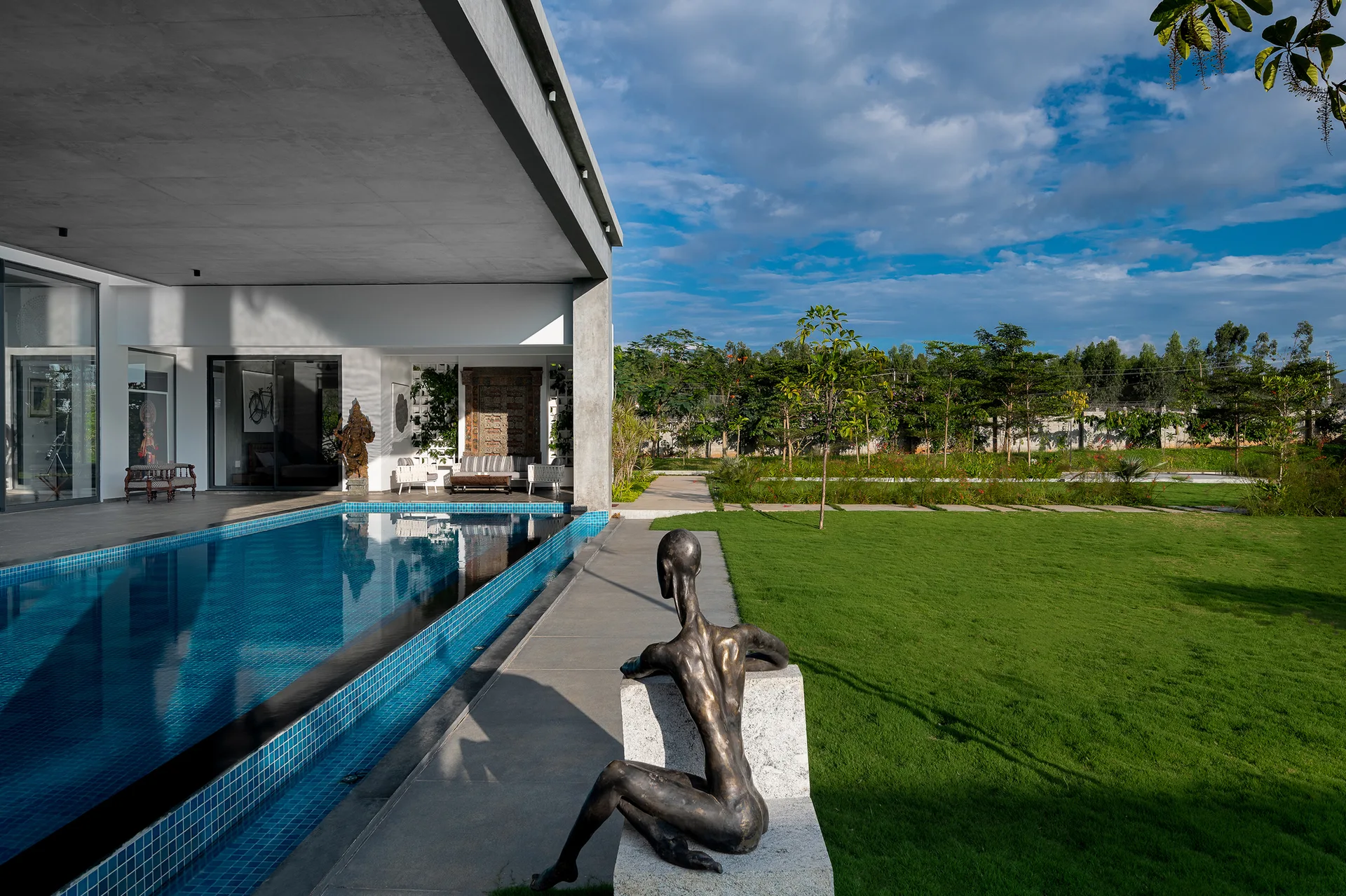 A modern concrete patio features a blue tiled swimming pool alongside an expansive green lawn with a bronze sculpture in the foreground.