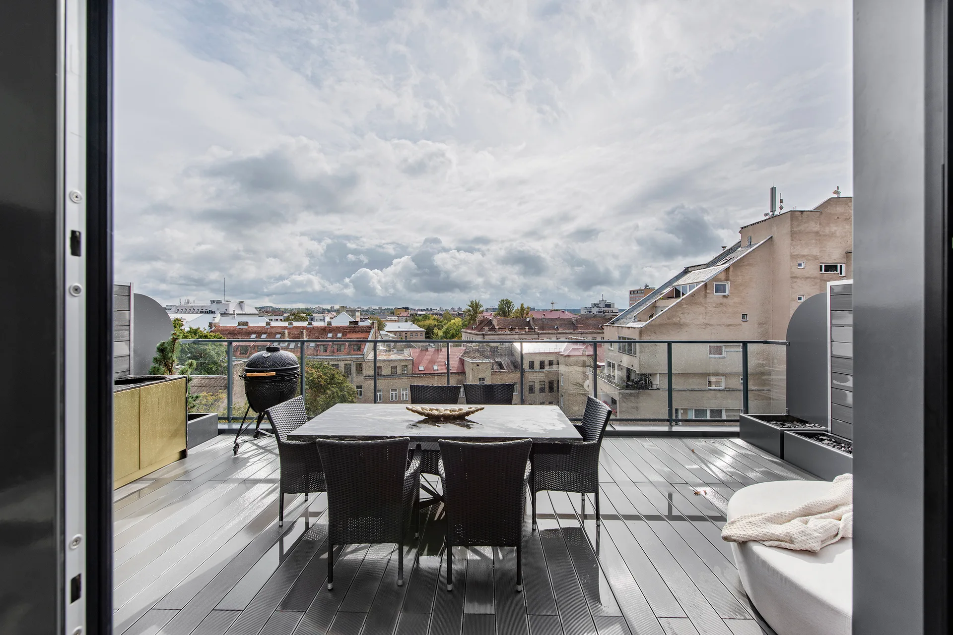 A rooftop terrace features a grey plank deck with a dark woven dining set, a black grill, a curved lounge chair, and a glass railing overlooking the city.
