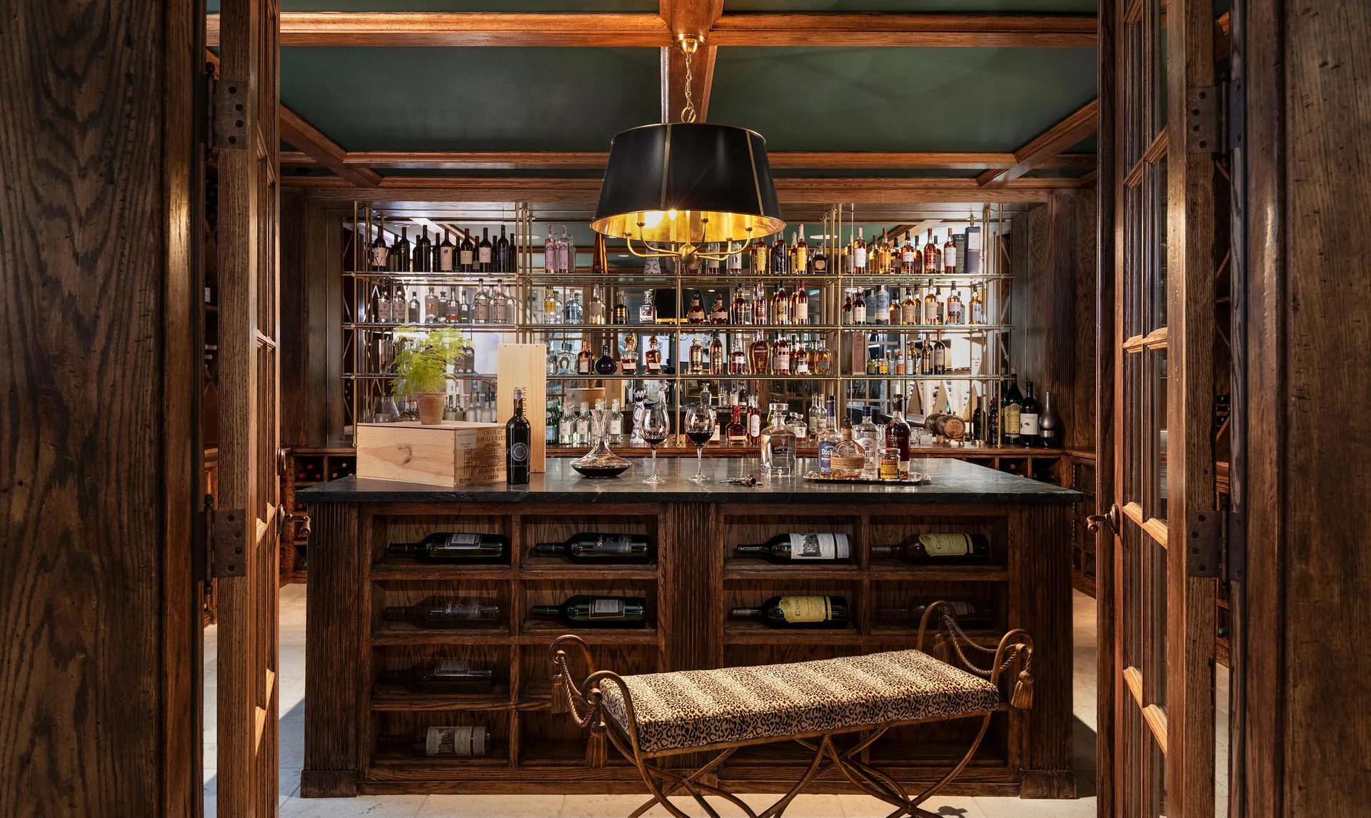 A traditional home bar features dark wood cabinetry, a stone-topped island, a leopard print bench, and a backlit shelving display.