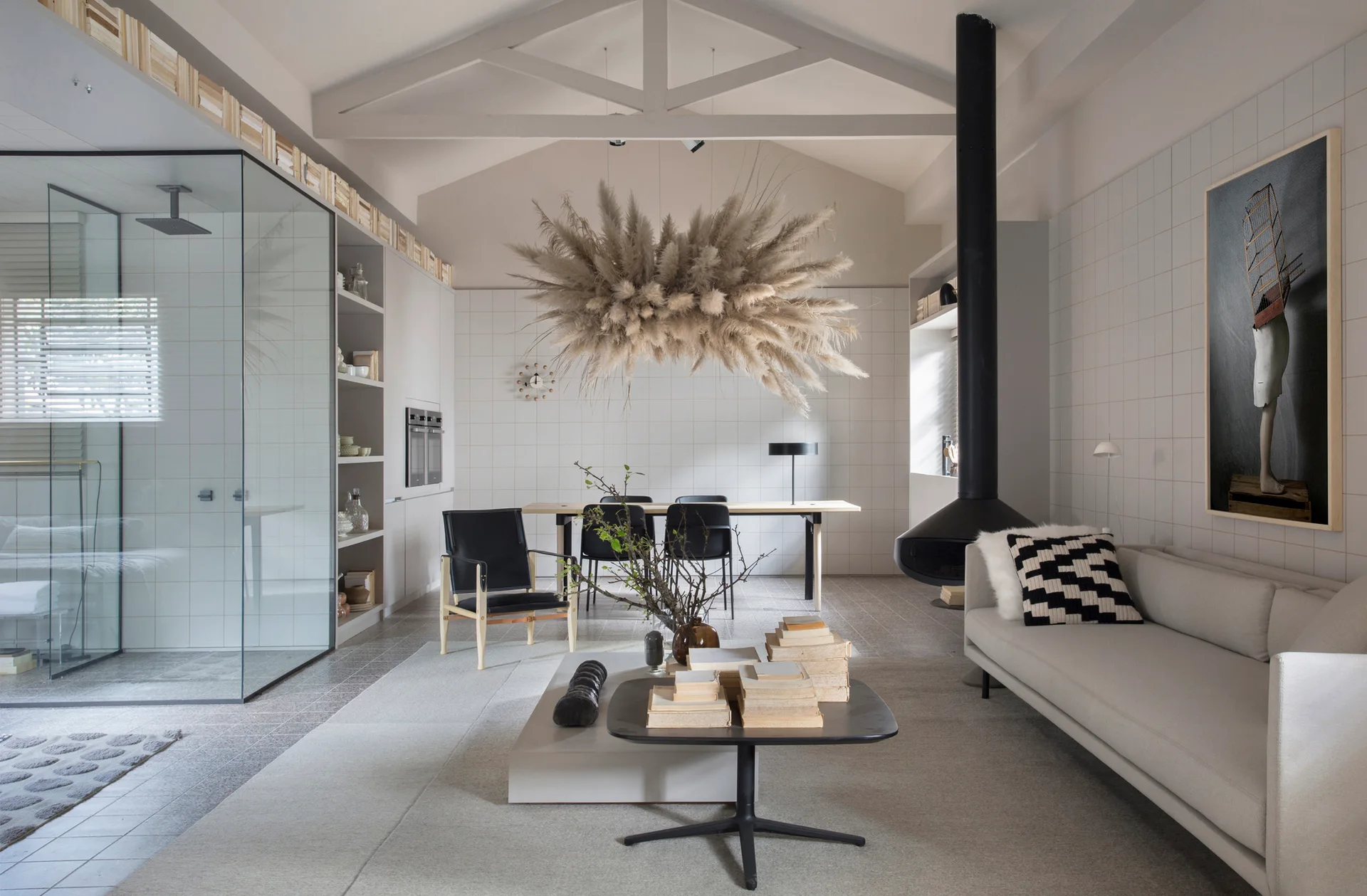 A minimalist open-plan living room featuring a suspended black fireplace, white grid tiled walls, and a hanging pampas grass installation.