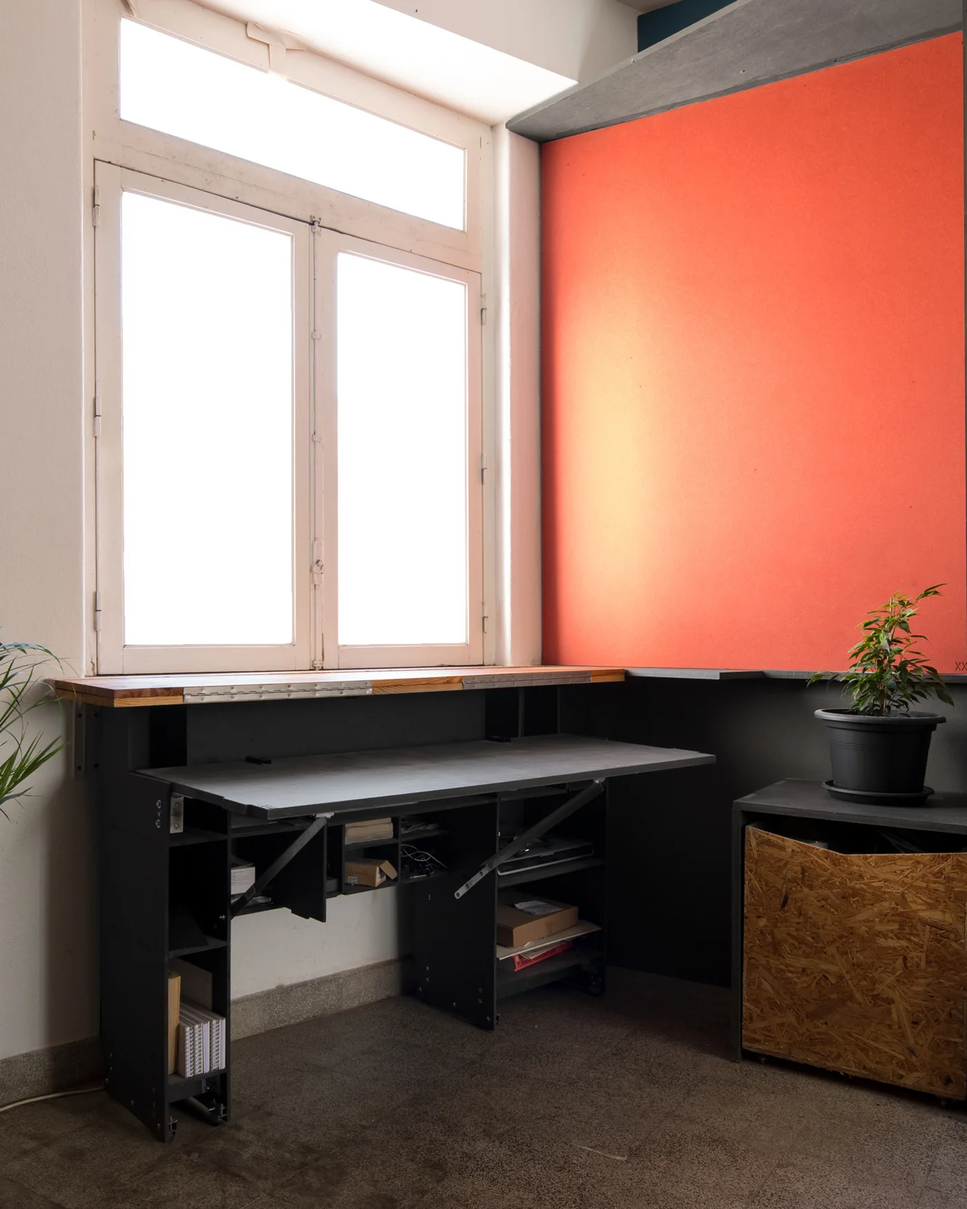 An industrial black desk sits under a large window next to an orange wall panel and an OSB cabinet.