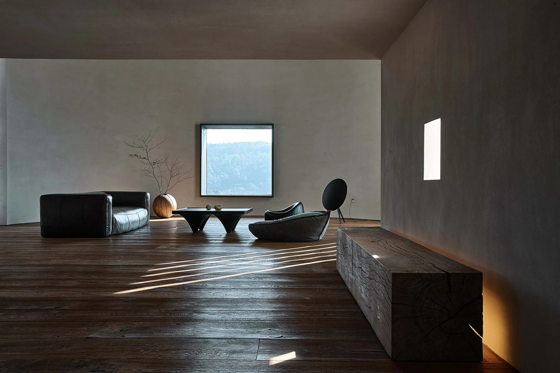 A minimalist living room features wide plank wood flooring, a low-profile leather sofa, a square window, and a rustic raw wood log bench.