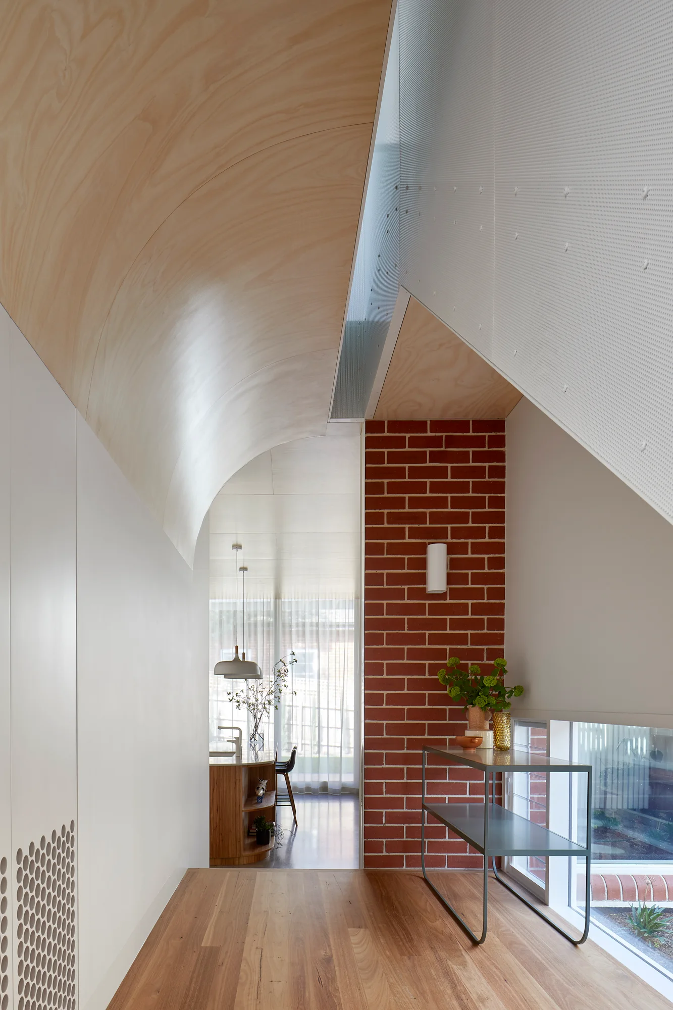 A modern hallway features a curved plywood ceiling, an exposed red brick wall, and a slim console table leading to a kitchen.