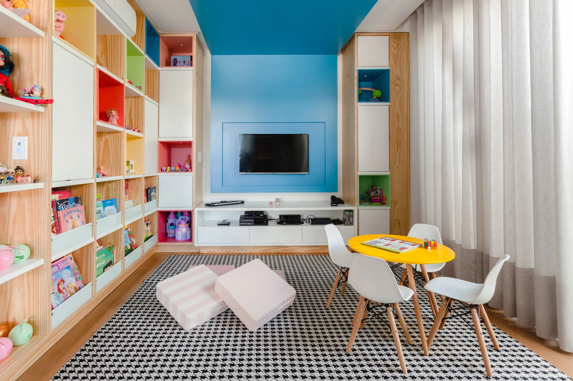 A playroom features a large plywood shelving unit with colorful cubbies, a yellow kids' table with chairs, and a blue accent wall with a mounted TV.