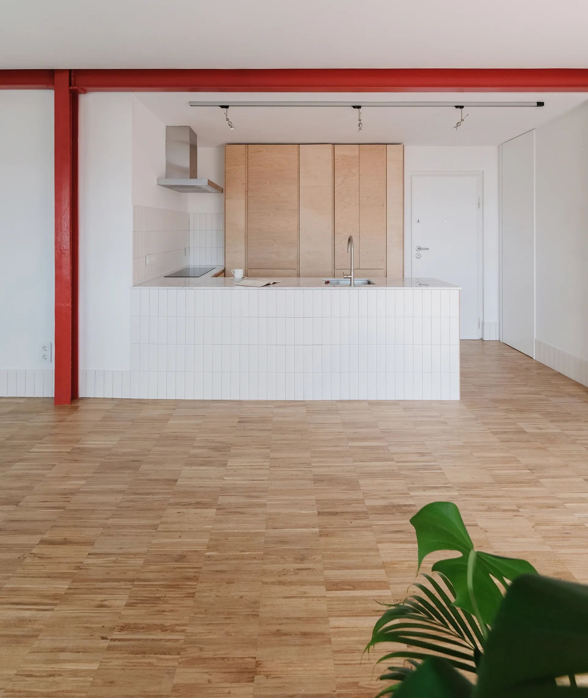 A minimalist kitchen features a white vertically tiled island, light wood cabinets, and a prominent red steel support beam on a parquet wood floor.