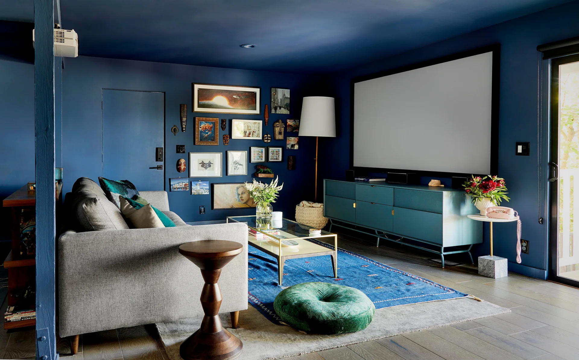 A media room with a grey sofa and a teal console beneath a large projection screen against a dark blue wall and ceiling.