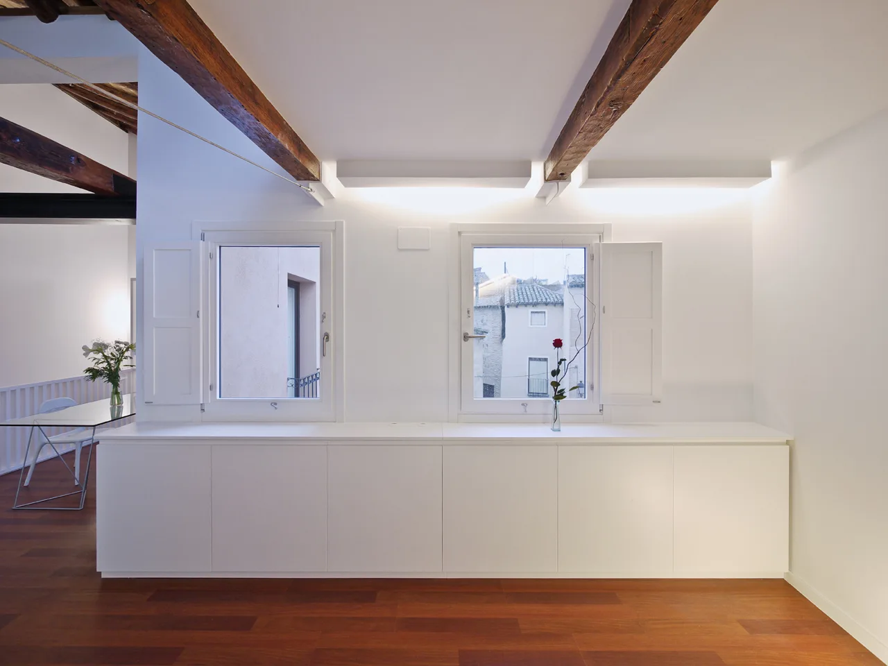 A minimalist white room features exposed dark wood ceiling beams, hardwood flooring, and a long seamless storage cabinet running beneath two shuttered windows.