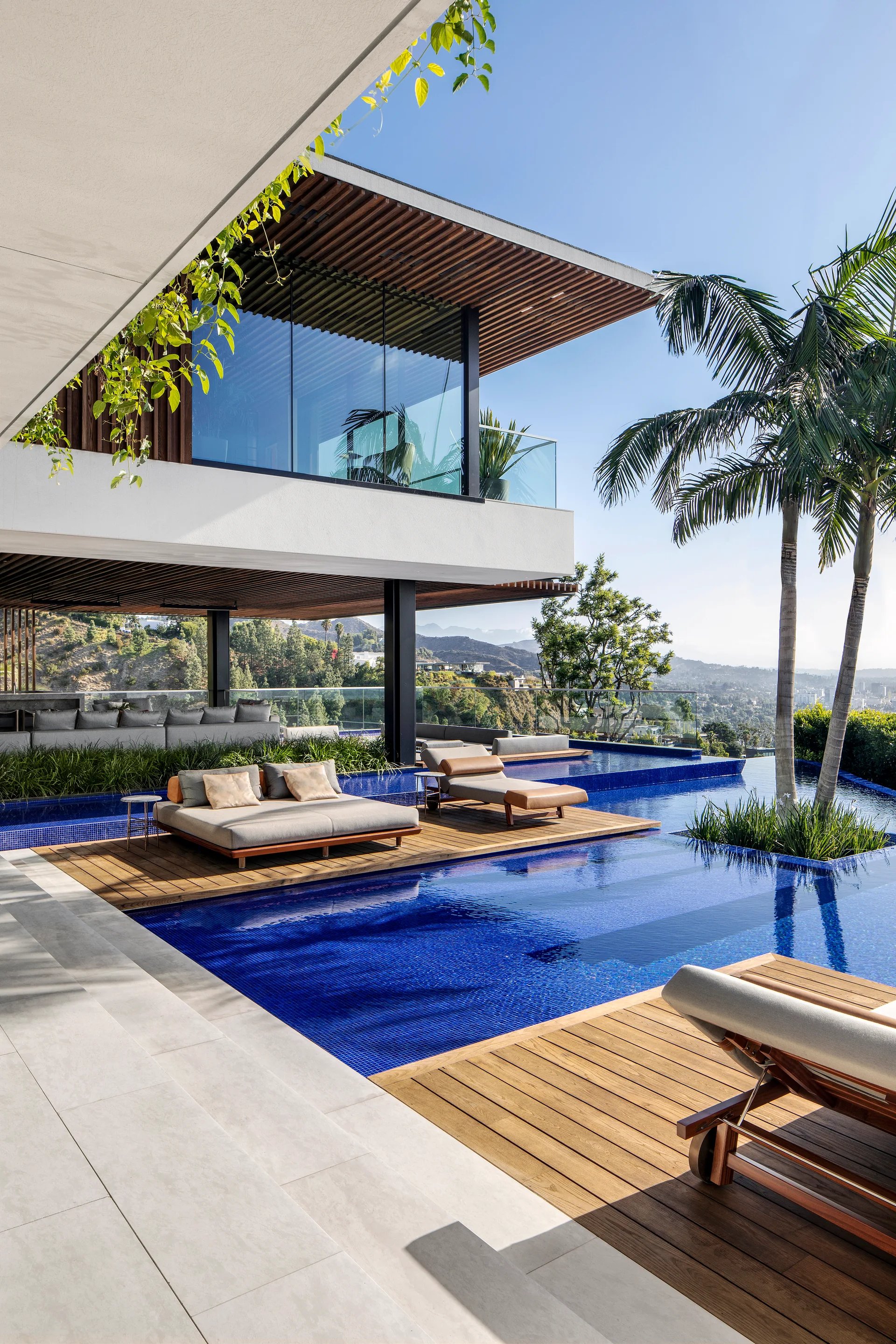 A modern outdoor pool area features blue tiled water, floating wooden deck platforms with lounge chairs, and palm trees against a hillside view.