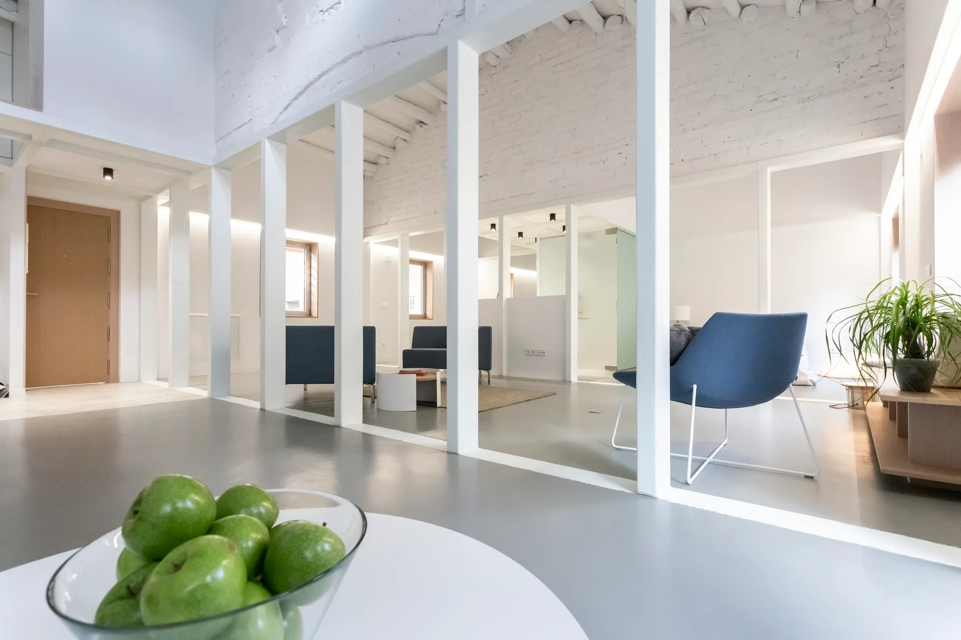 A minimalist interior features a series of white structural pillars separating seating areas with blue armchairs against a white painted brick wall.