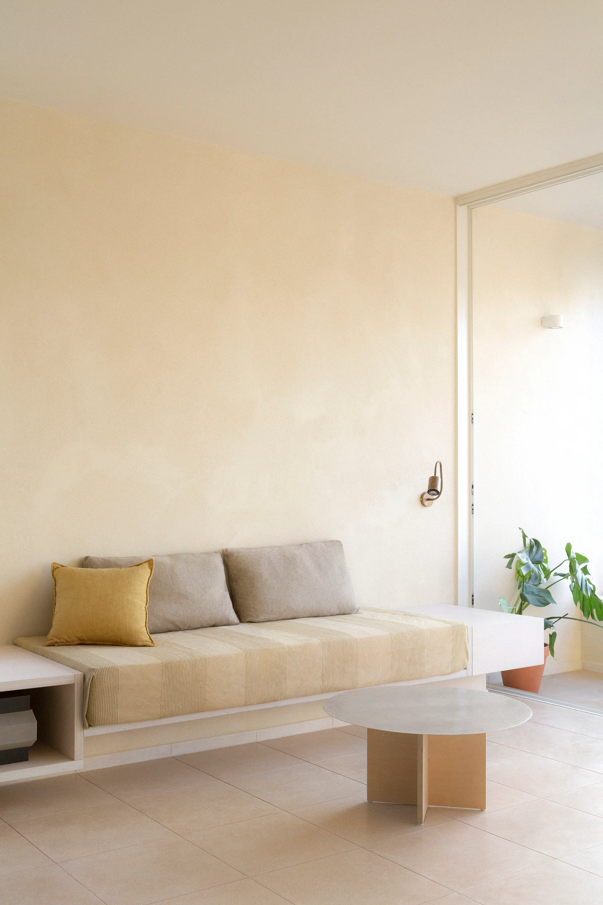 A minimalist living space features a built-in daybed with beige cushions, a low round coffee table, and warm beige plaster walls.