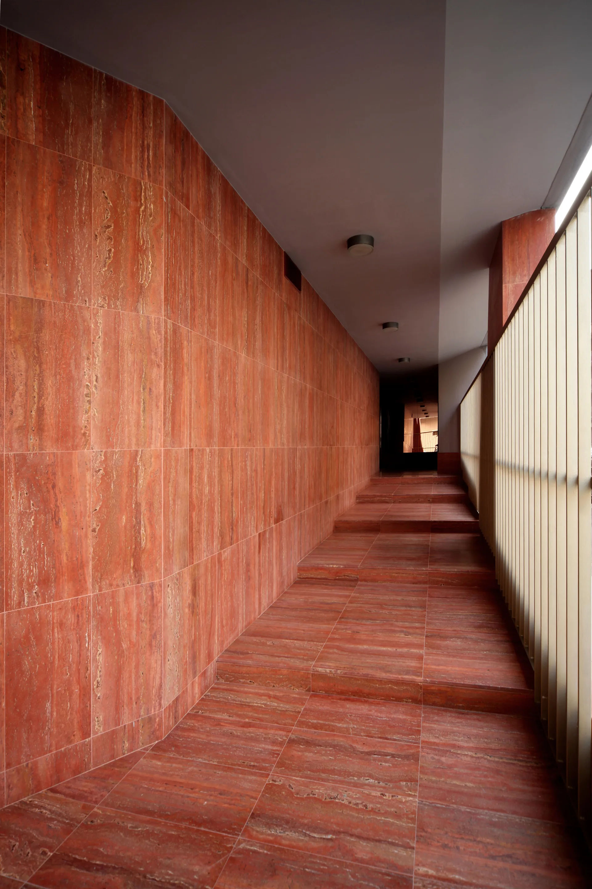 A long interior hallway with polished red stone clad walls and stepped floors, bordered by vertical metal railings on the right.