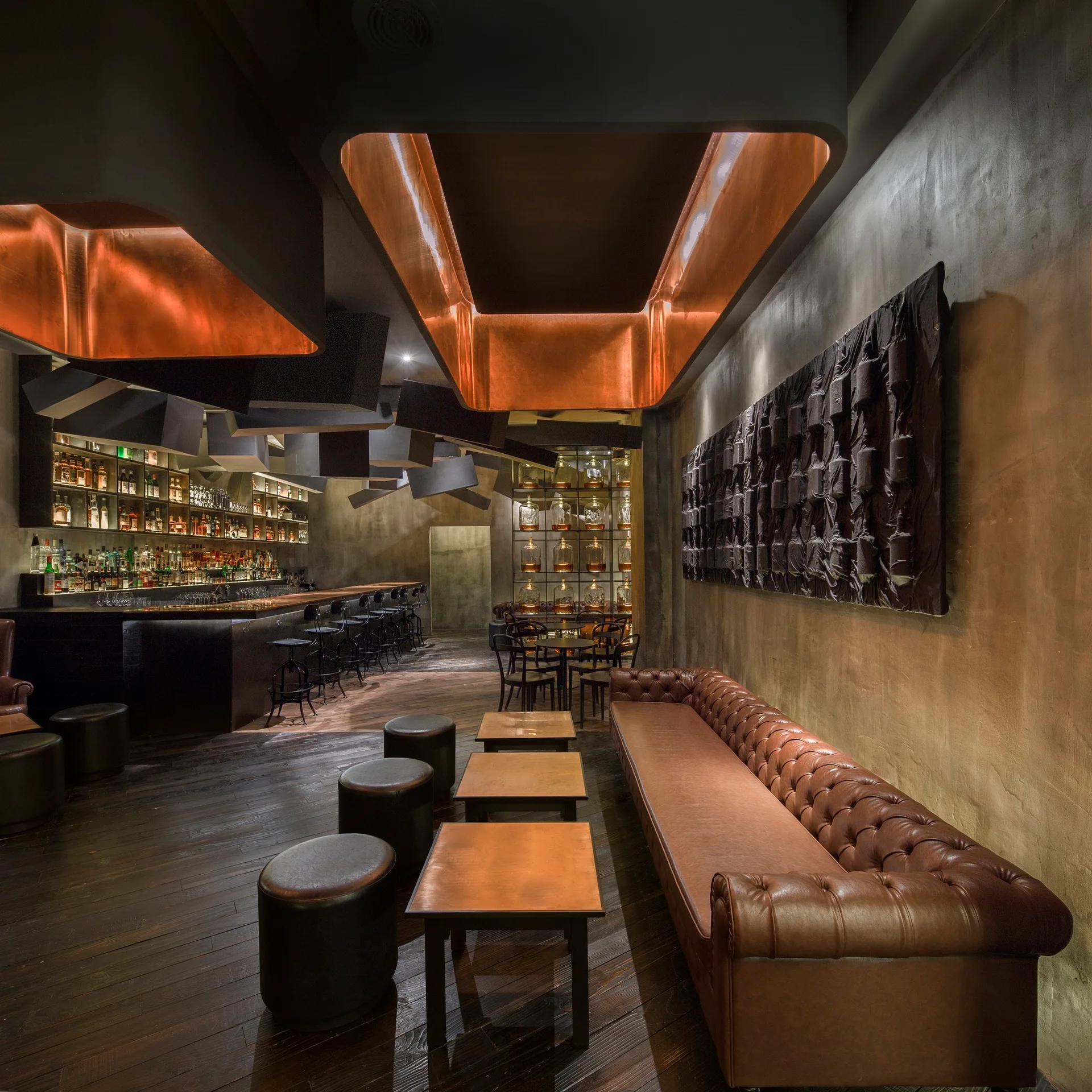 An atmospheric dark bar interior featuring a long tufted leather sofa, copper-topped tables, and glowing copper-lined ceiling light fixtures.