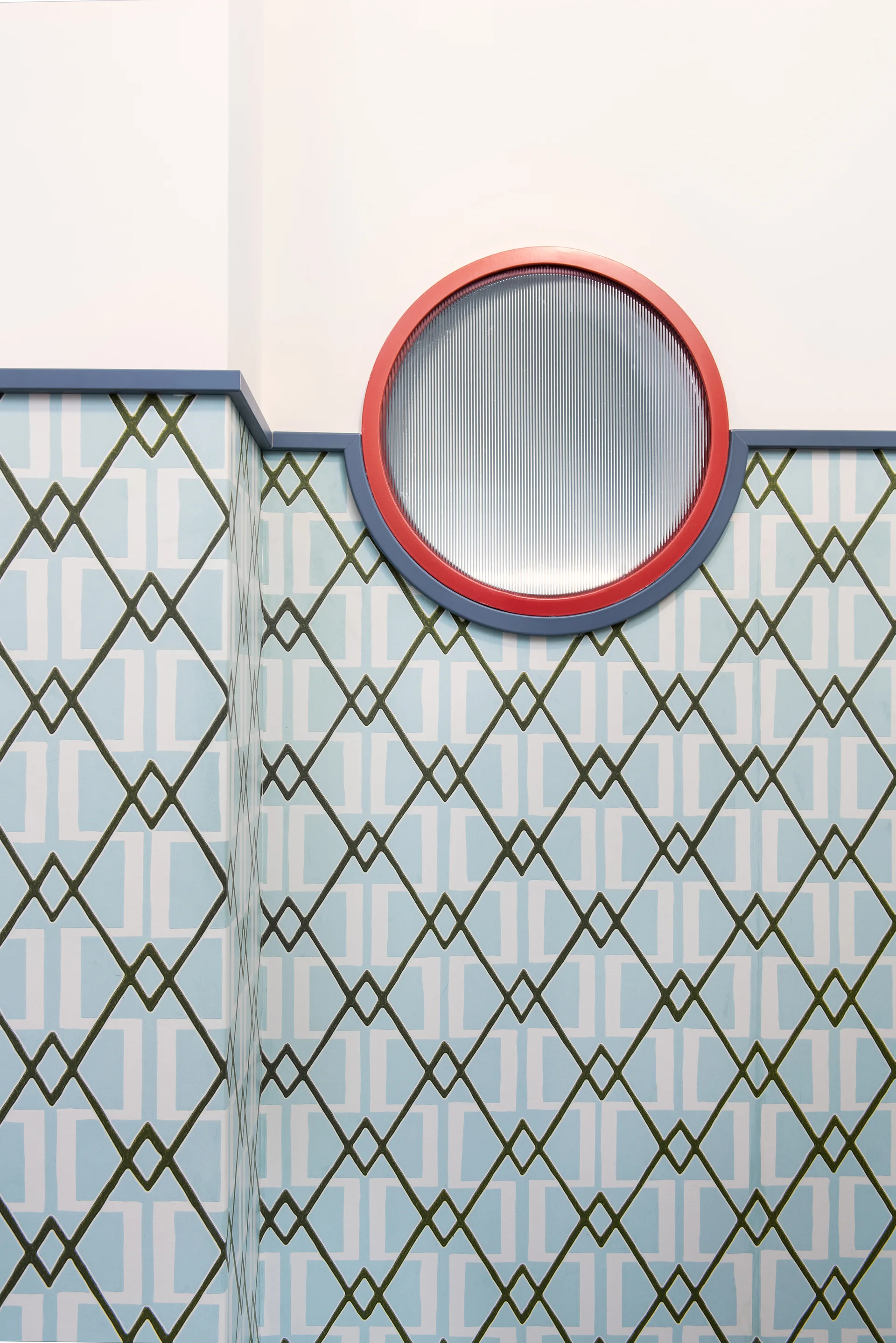 A circular window with reeded glass and a bright red frame sits above blue-and-white geometric wallpaper and coordinating blue trim.