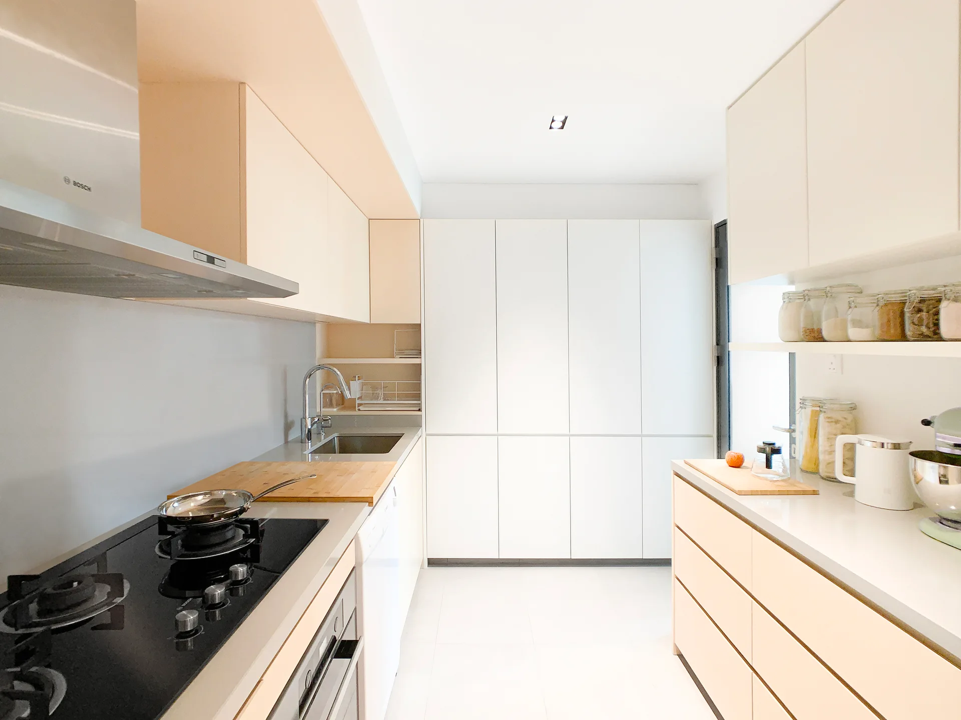 A minimalist galley kitchen features off-white flat-front cabinetry, a black cooktop with a stainless steel range hood, and light-colored floor tiles.