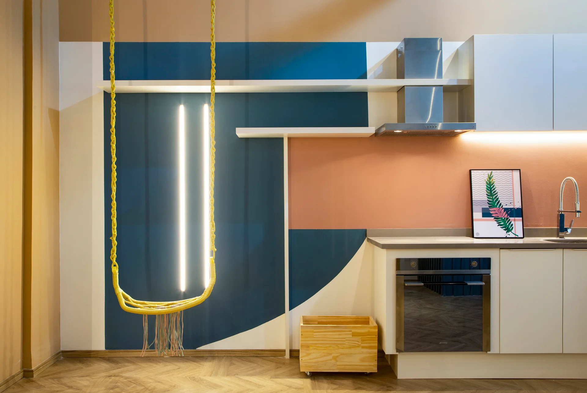 A modern kitchen features white cabinetry, color-blocked walls in blue and peach, herringbone wood floors, and a yellow hanging swing.