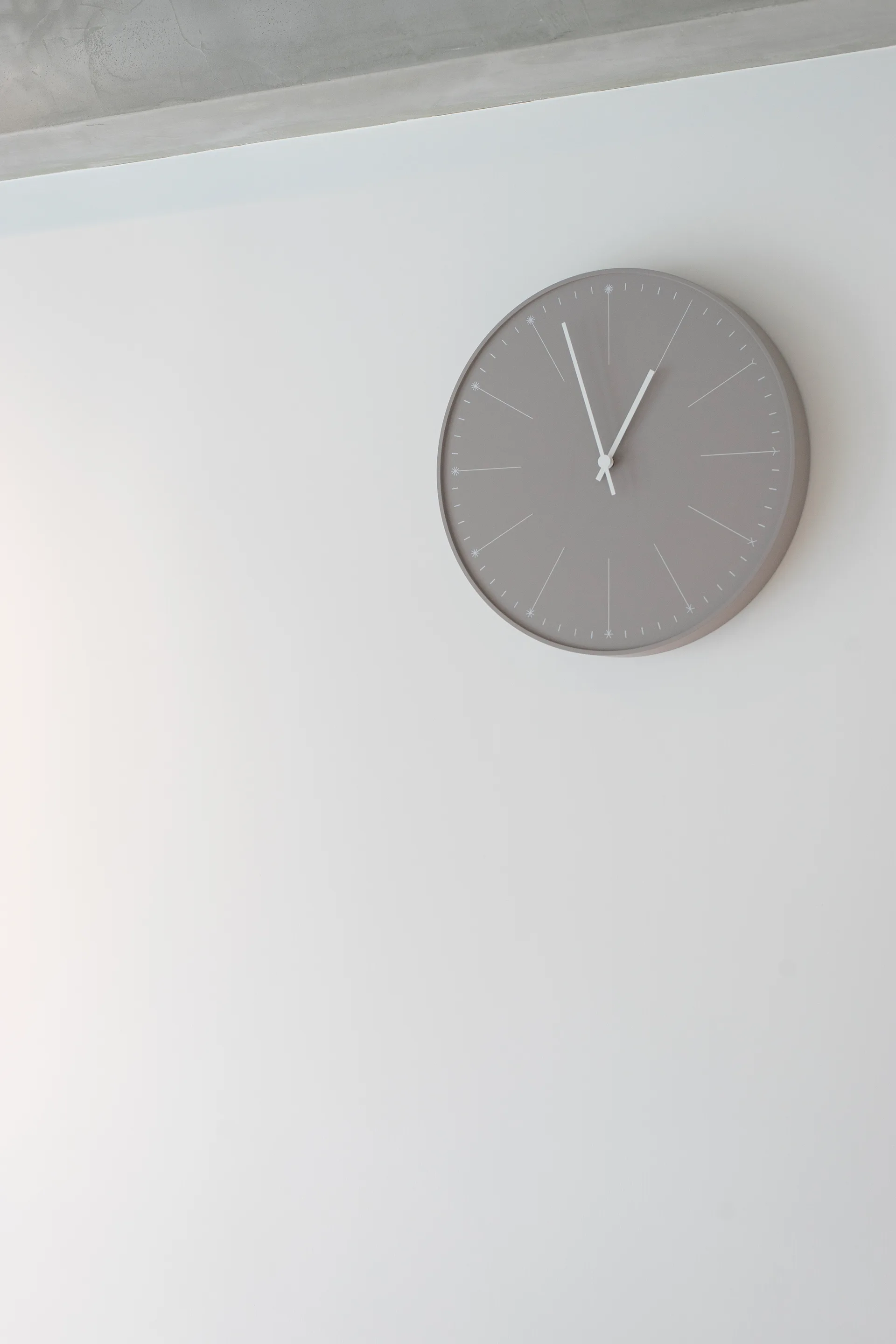 A round gray wall clock with slender white hands hangs on a plain white wall below an exposed concrete ceiling beam.