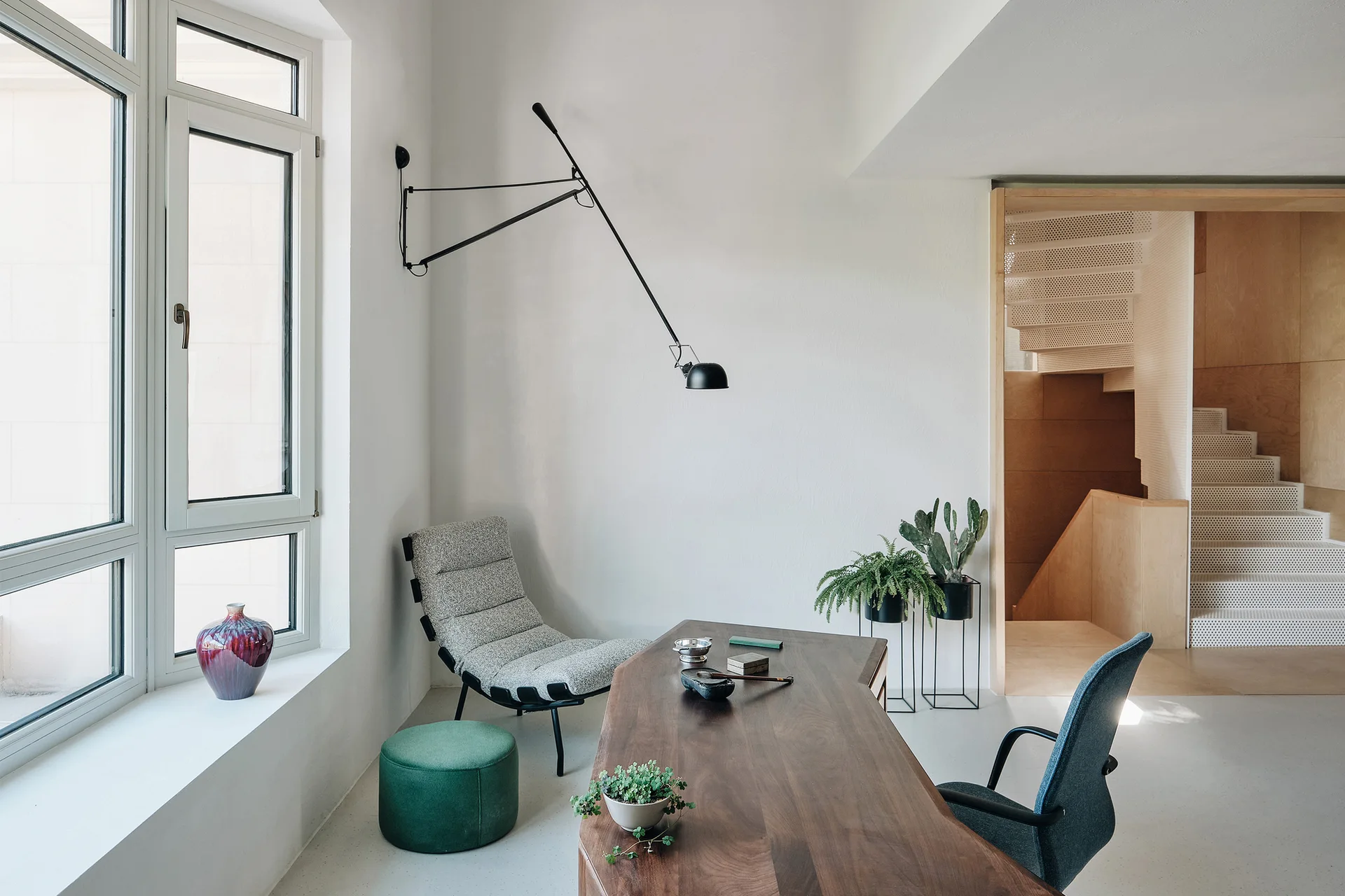 A minimalist home office featuring a wooden desk, a grey lounge chair, a green ottoman, and a perforated metal staircase in the background.