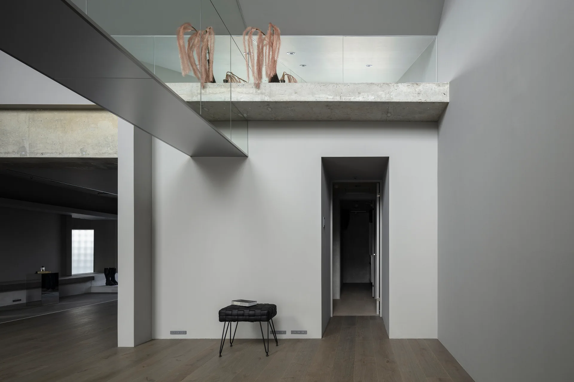 A minimalist gray hallway features a suspended metal bridge, a concrete mezzanine with a glass railing, and a small black tufted ottoman on wood flooring.